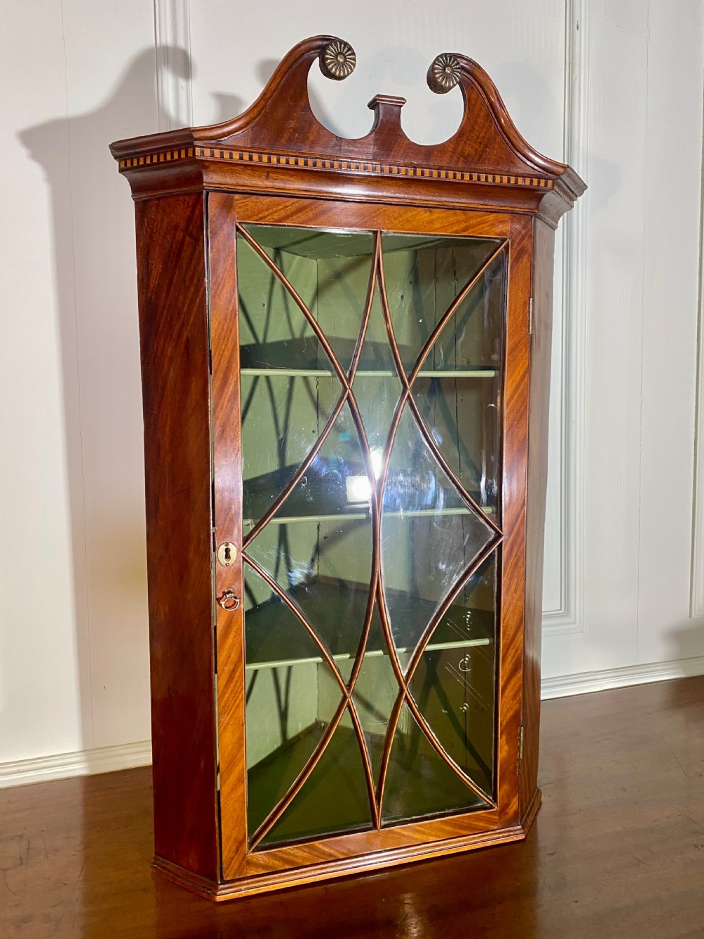 An eighteenth-century mahogany glazed corner cupboard with a swan neck pediment, carved rosettes, dentil inlay, and a green painted interior.