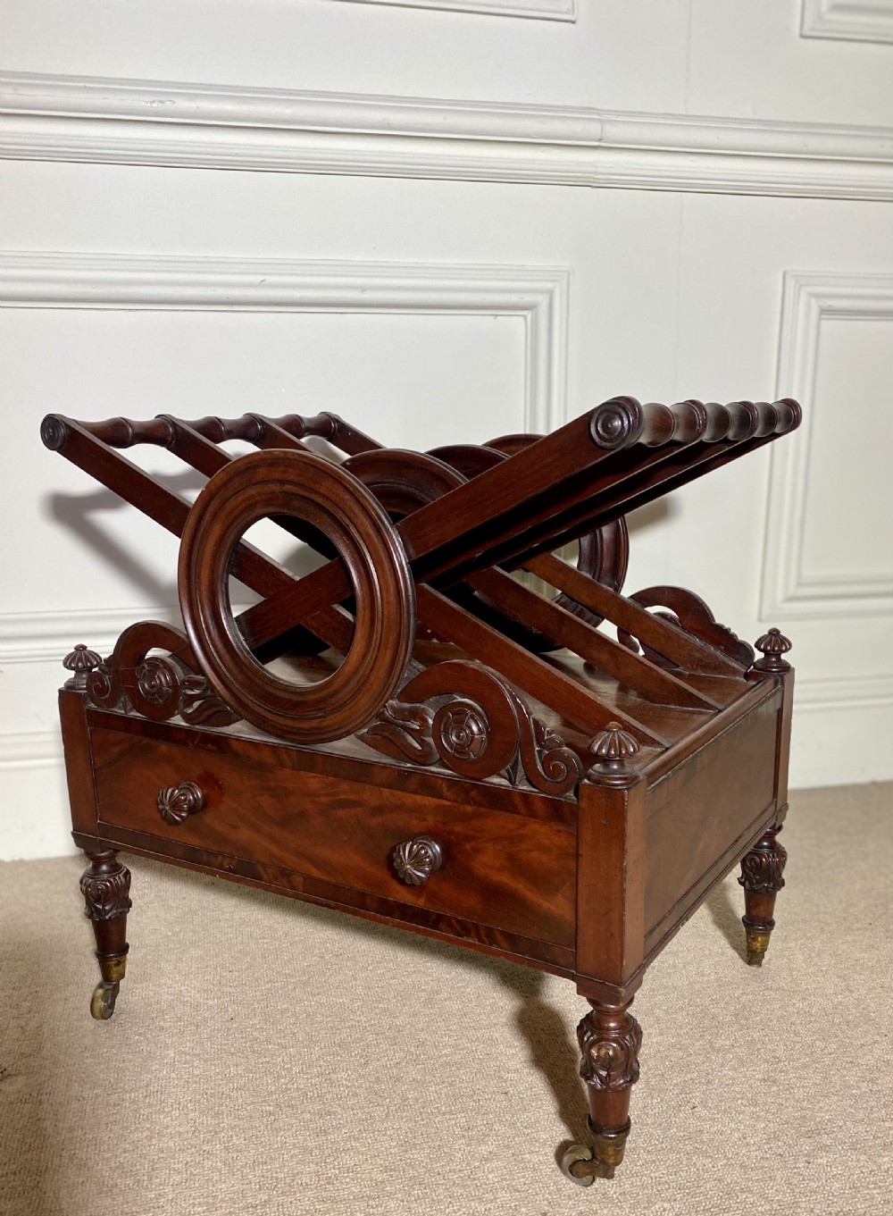A Regency mahogany music canterbury featuring circular divided compartments, carved rosettes, a lower drawer with turned knobs, and carved legs on brass castors. View V