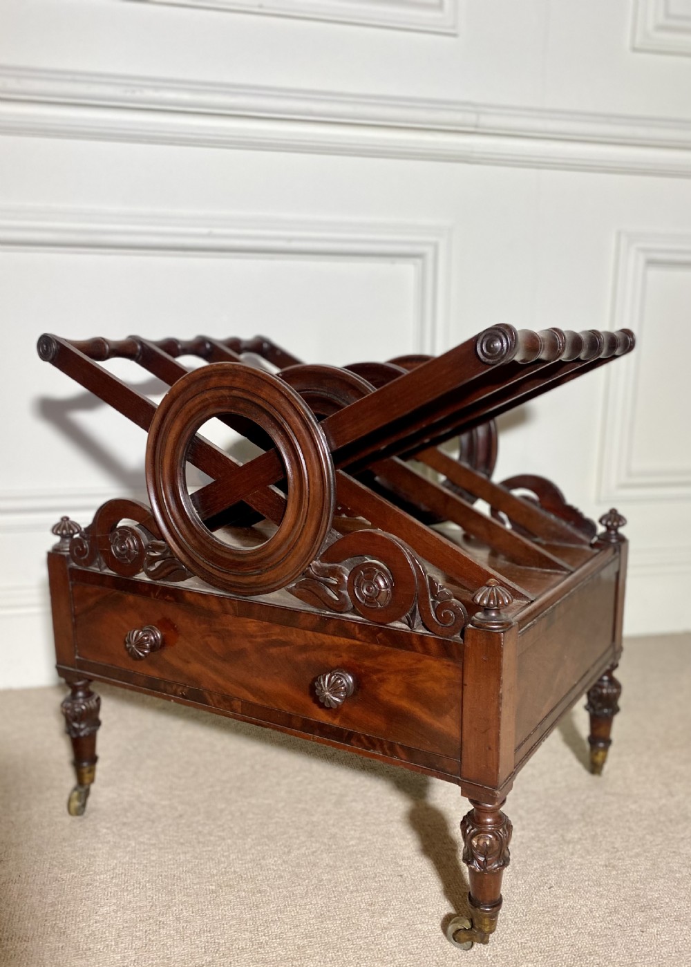 A Regency mahogany music canterbury featuring circular divided compartments, carved rosettes, a lower drawer with turned knobs, and carved legs on brass castors. View VII