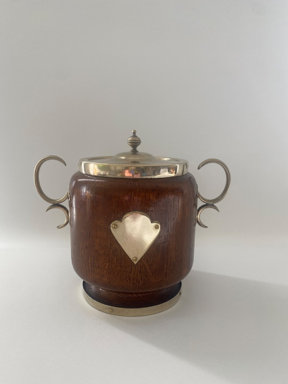 A turned oak ice bucket featuring silver-plated scroll handles, a shield-shaped front cartouche, and a domed metal lid with a finial.