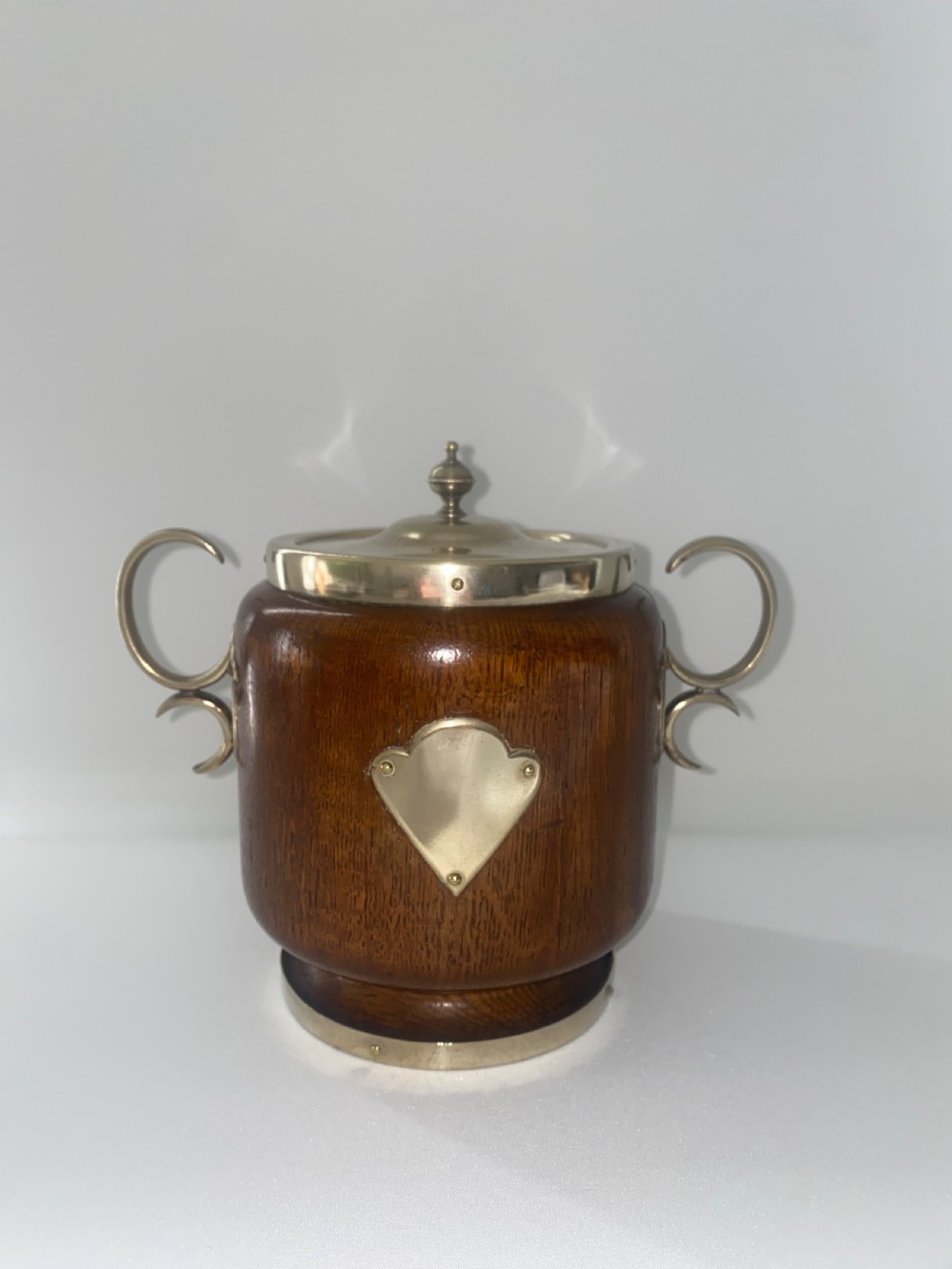 A turned oak ice bucket featuring silver-plated scroll handles, a shield-shaped front cartouche, and a domed metal lid with a finial. View II