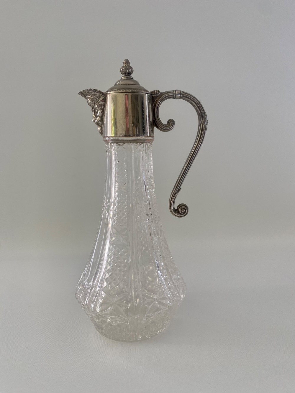 A cut crystal claret jug featuring a silver-plated collar, a mascaron face spout, and an ornate scroll handle.
