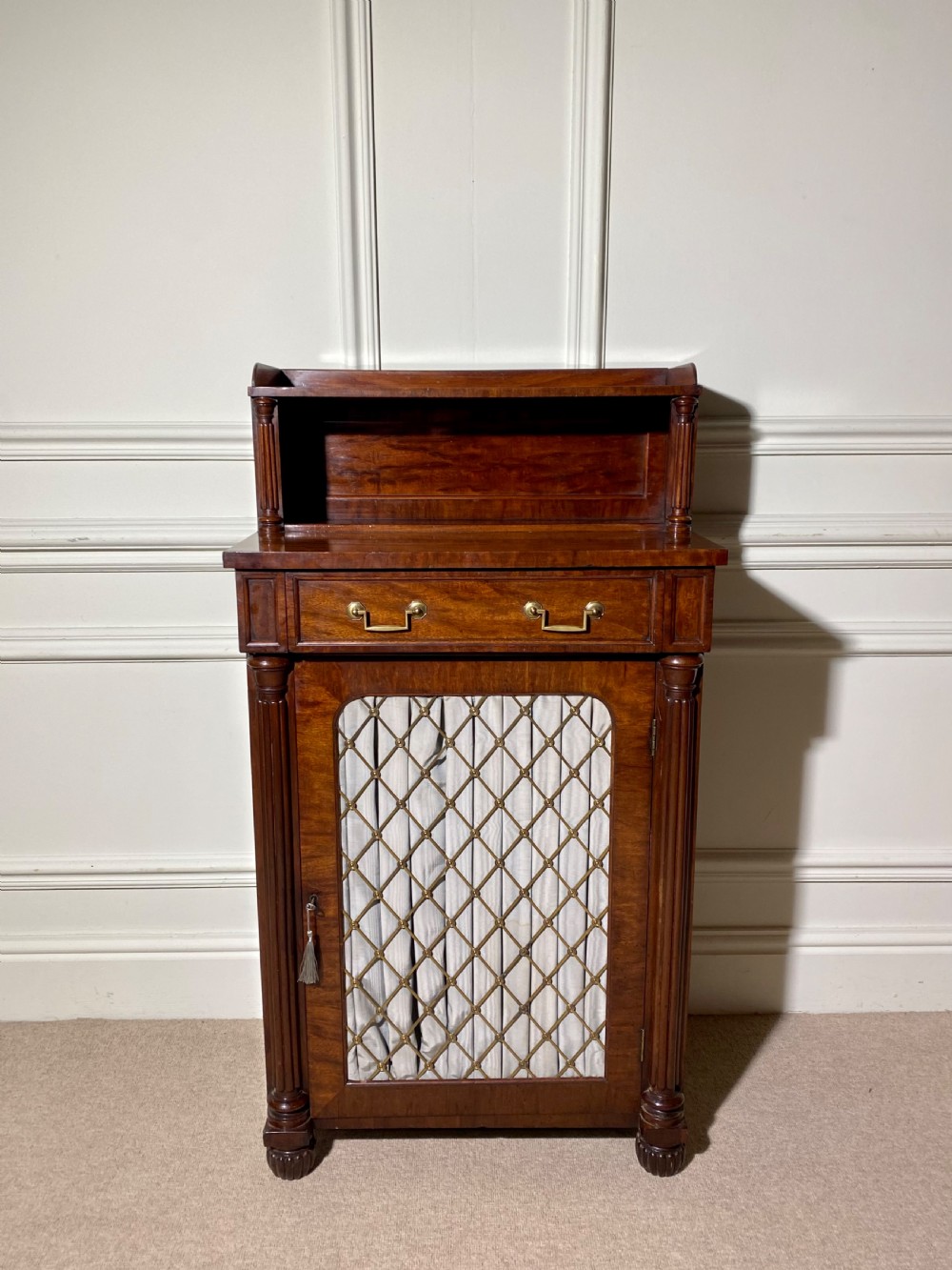 A Regency mahogany chiffonier featuring a raised shelf with reeded column supports above a frieze drawer and a lower cupboard door with brass wire grille and pleated fabric.