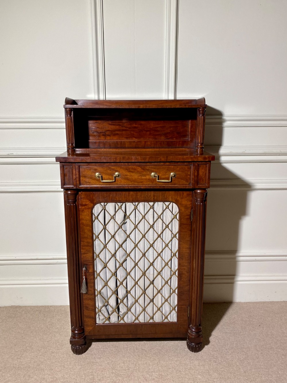 A Regency mahogany chiffonier featuring a raised shelf with reeded column supports above a frieze drawer and a lower cupboard door with brass wire grille and pleated fabric. View II