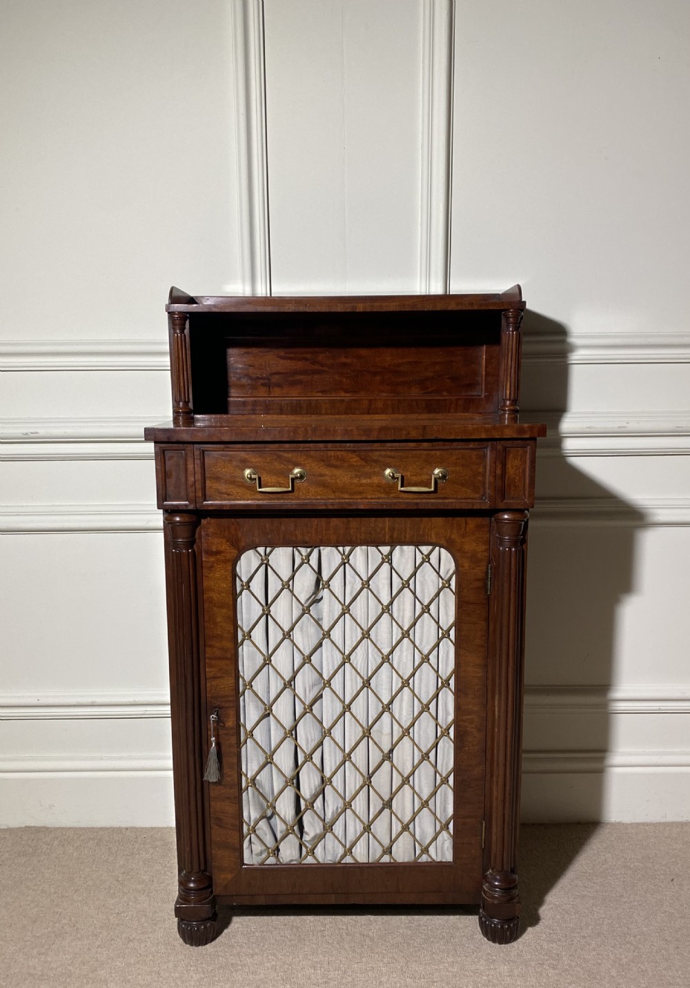 A Regency mahogany chiffonier featuring a raised shelf with reeded column supports above a frieze drawer and a lower cupboard door with brass wire grille and pleated fabric. View III
