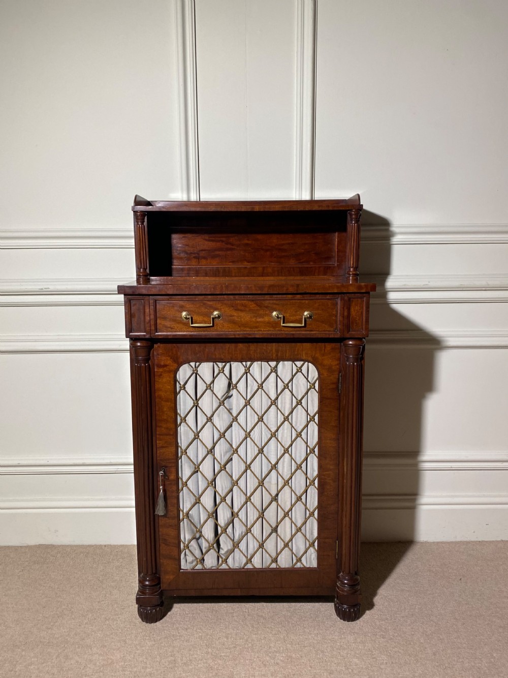 A Regency mahogany chiffonier featuring a raised shelf with reeded column supports above a frieze drawer and a lower cupboard door with brass wire grille and pleated fabric. View VI