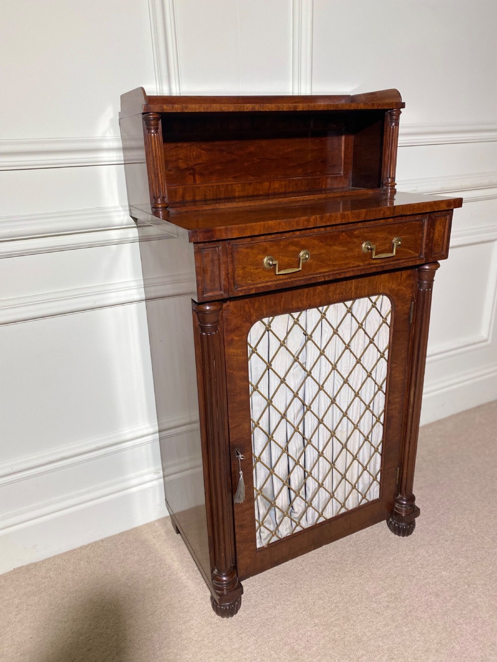 A Regency mahogany chiffonier featuring a raised shelf with reeded column supports above a frieze drawer and a lower cupboard door with brass wire grille and pleated fabric. View VII