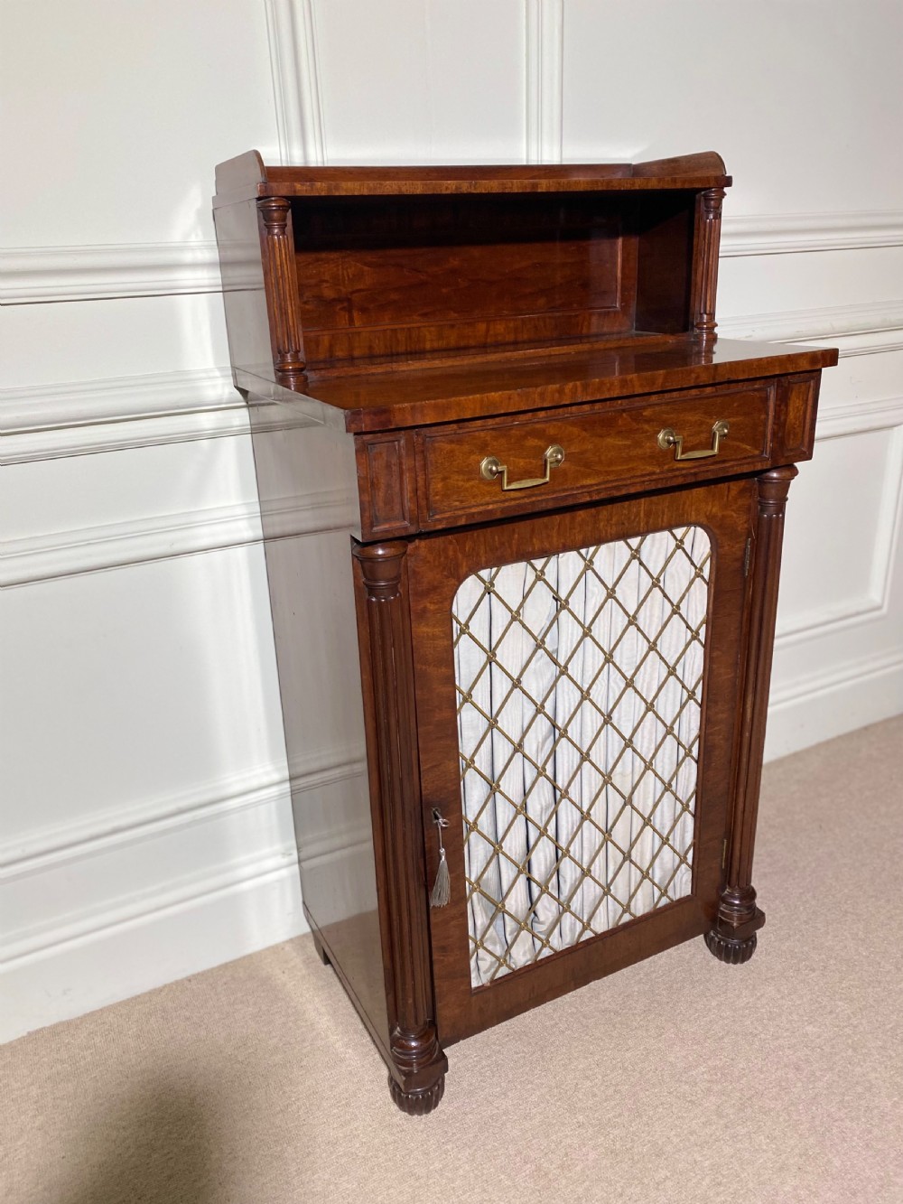 A Regency mahogany chiffonier featuring a raised shelf with reeded column supports above a frieze drawer and a lower cupboard door with brass wire grille and pleated fabric. View VIII