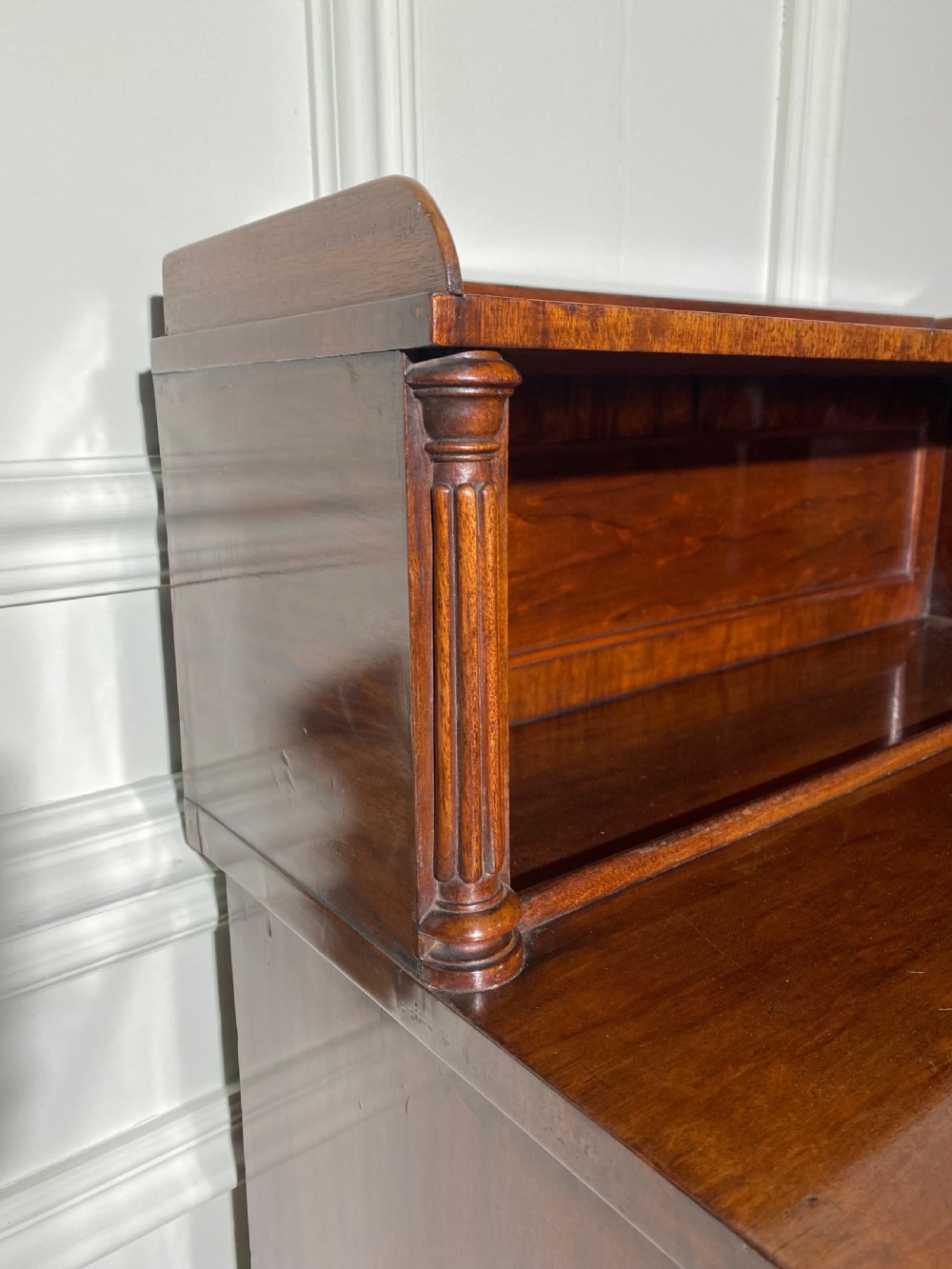 A Regency mahogany chiffonier featuring a raised shelf with reeded column supports above a frieze drawer and a lower cupboard door with brass wire grille and pleated fabric. View IX