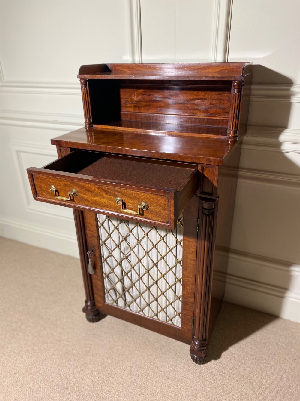 A Regency mahogany chiffonier featuring a raised shelf with reeded column supports above a frieze drawer and a lower cupboard door with brass wire grille and pleated fabric. View XI