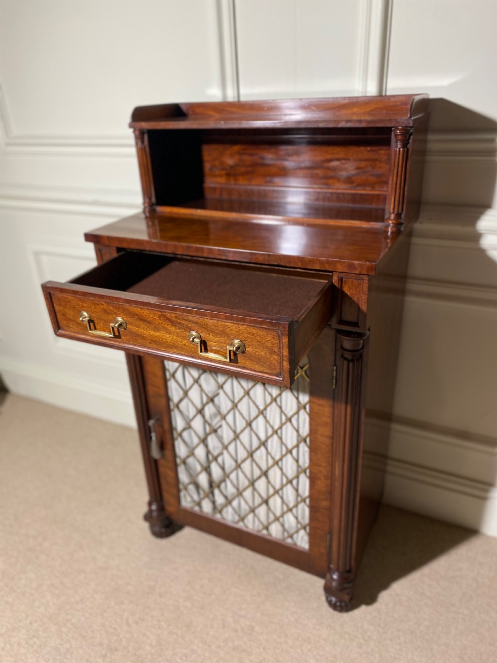 A Regency mahogany chiffonier featuring a raised shelf with reeded column supports above a frieze drawer and a lower cupboard door with brass wire grille and pleated fabric. View XII