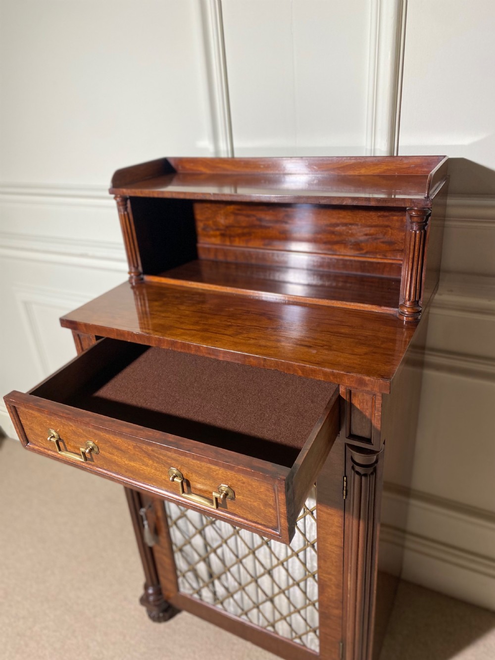 A Regency mahogany chiffonier featuring a raised shelf with reeded column supports above a frieze drawer and a lower cupboard door with brass wire grille and pleated fabric. View XIII
