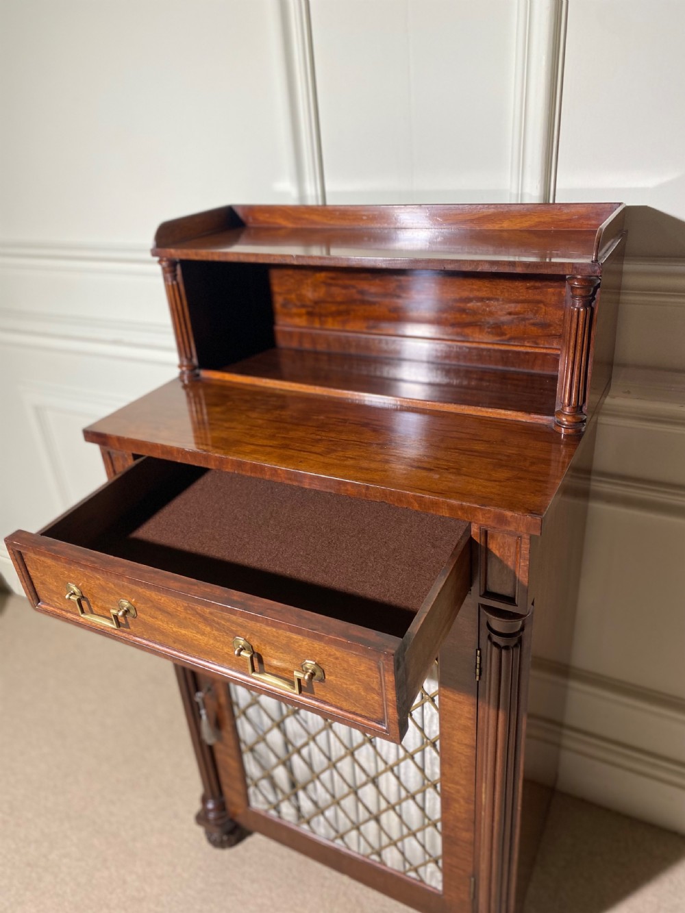 A Regency mahogany chiffonier featuring a raised shelf with reeded column supports above a frieze drawer and a lower cupboard door with brass wire grille and pleated fabric. View XIV