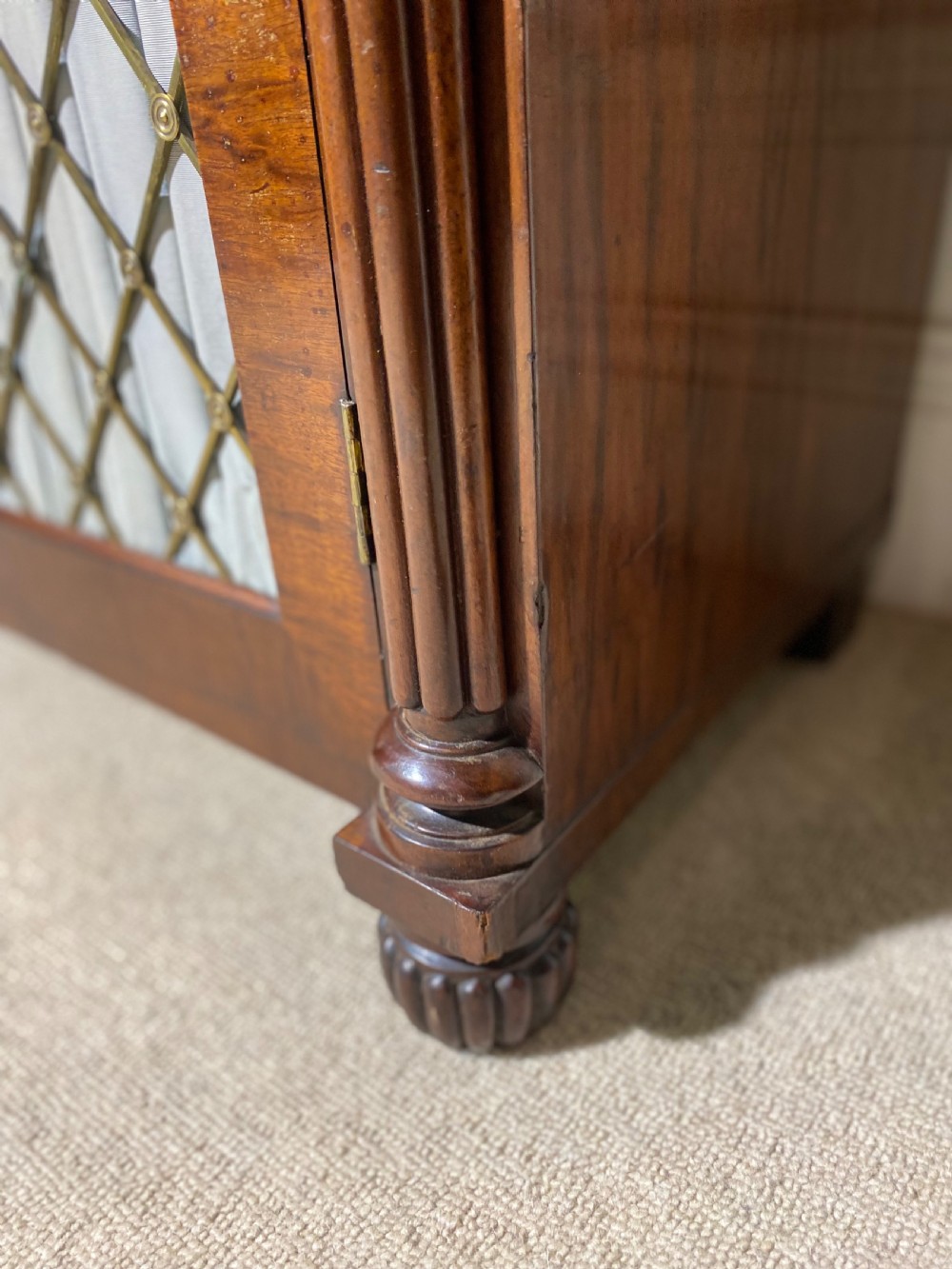 A Regency mahogany chiffonier featuring a raised shelf with reeded column supports above a frieze drawer and a lower cupboard door with brass wire grille and pleated fabric. View XV