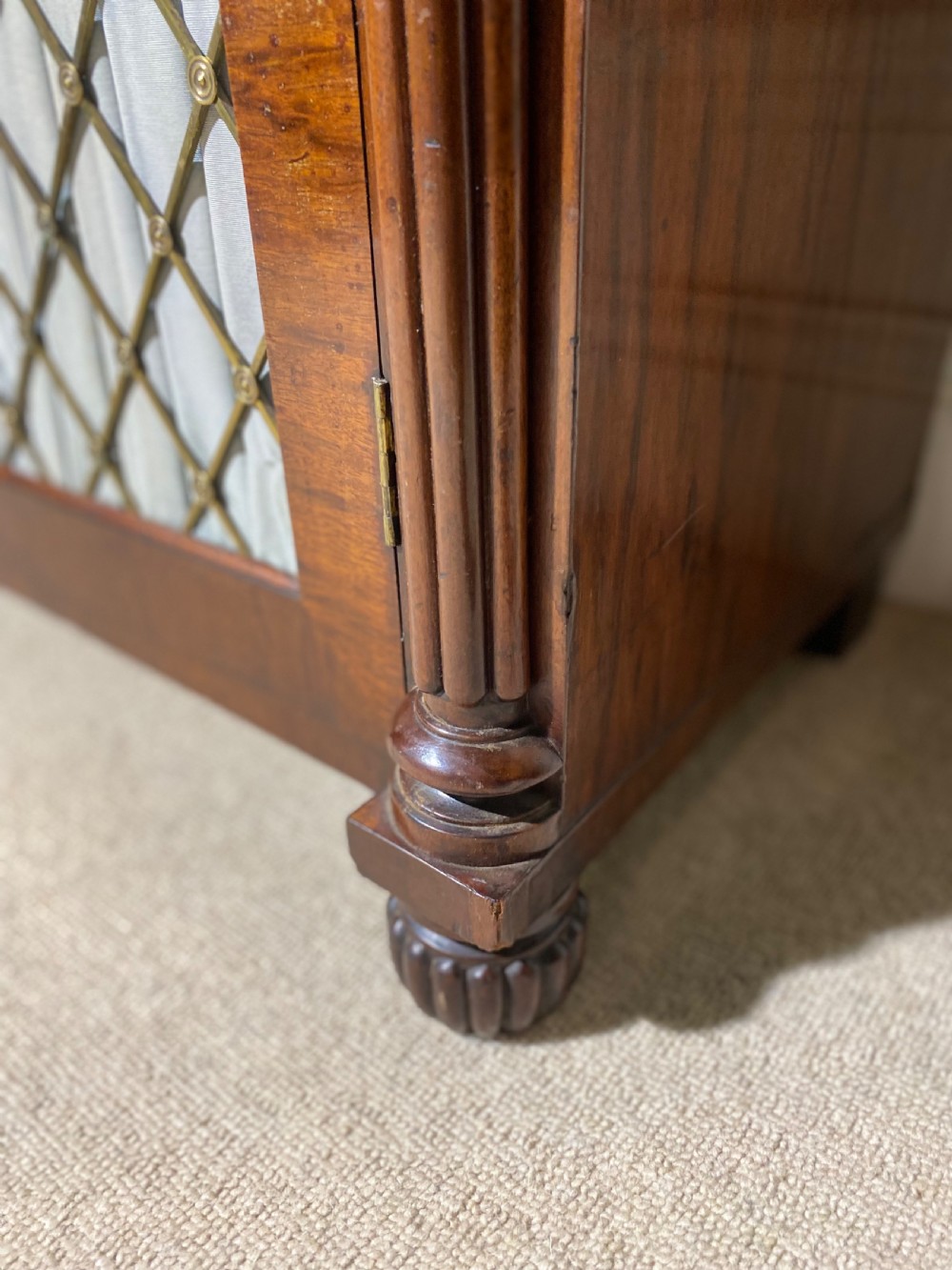 A Regency mahogany chiffonier featuring a raised shelf with reeded column supports above a frieze drawer and a lower cupboard door with brass wire grille and pleated fabric. View XVI