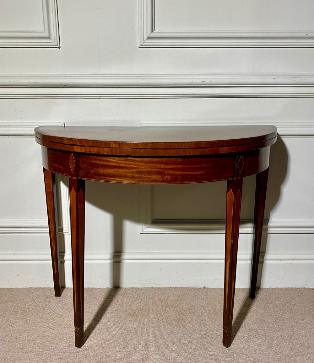An 18th-century Sheraton mahogany demi-lune tea table with line inlay and tapered square legs against a white wall.