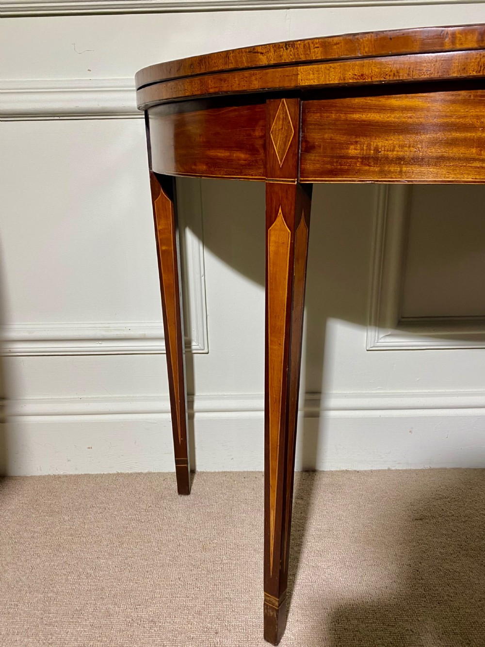 An 18th-century Sheraton mahogany demi-lune tea table with line inlay and tapered square legs against a white wall. View III