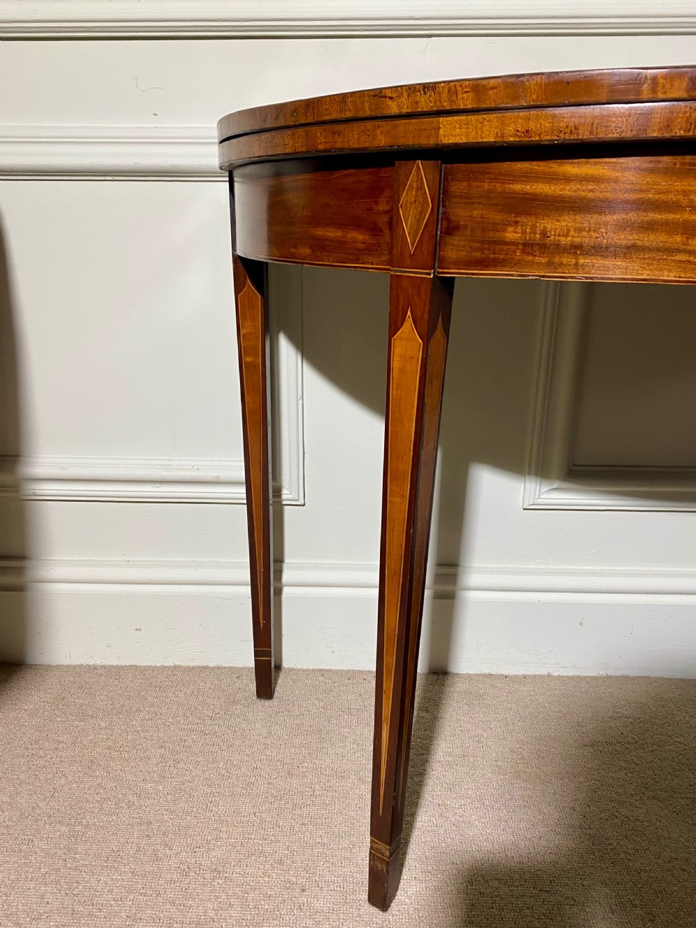 An 18th-century Sheraton mahogany demi-lune tea table with line inlay and tapered square legs against a white wall. View V
