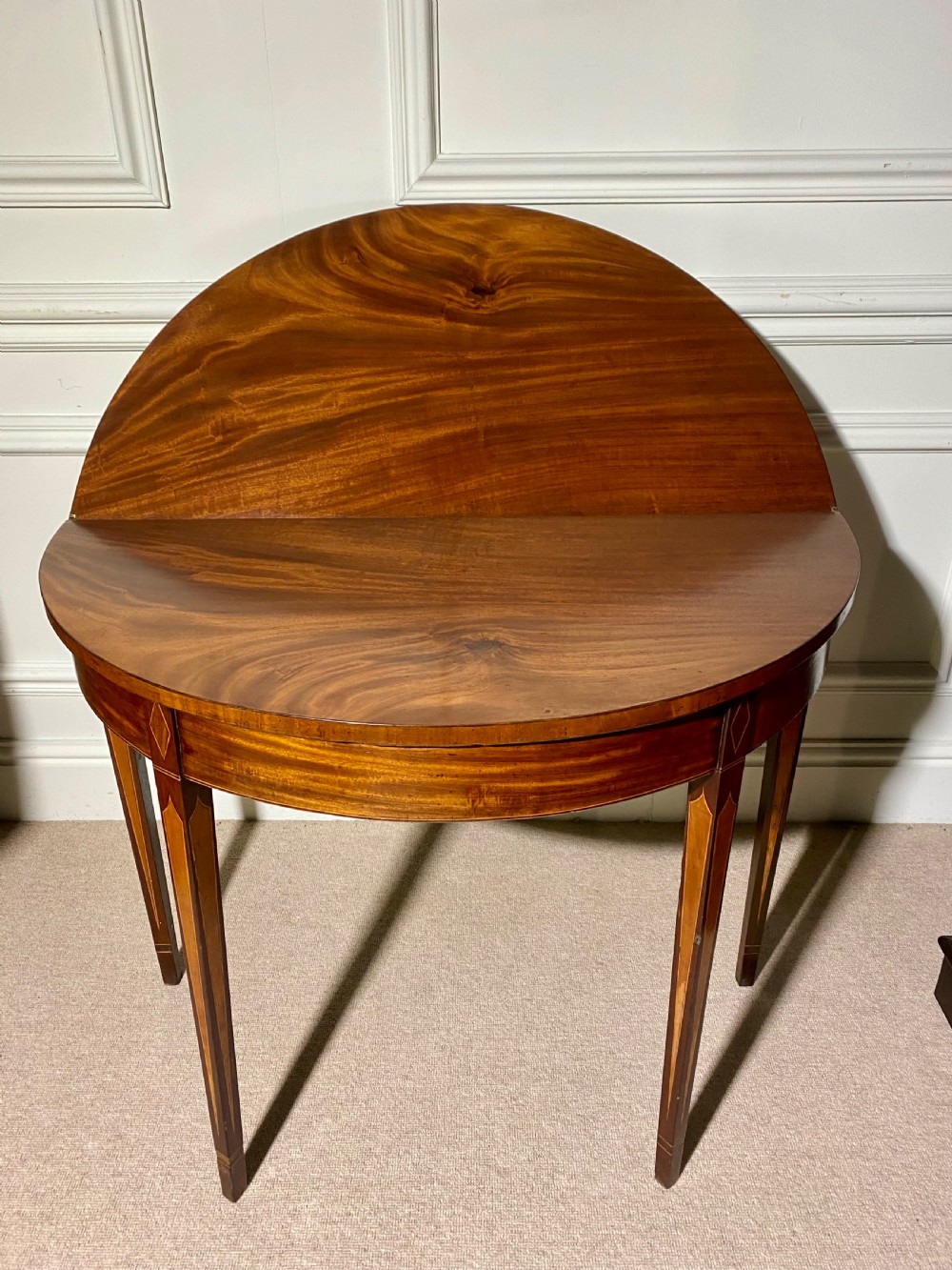 An 18th-century Sheraton mahogany demi-lune tea table with line inlay and tapered square legs against a white wall. View VII