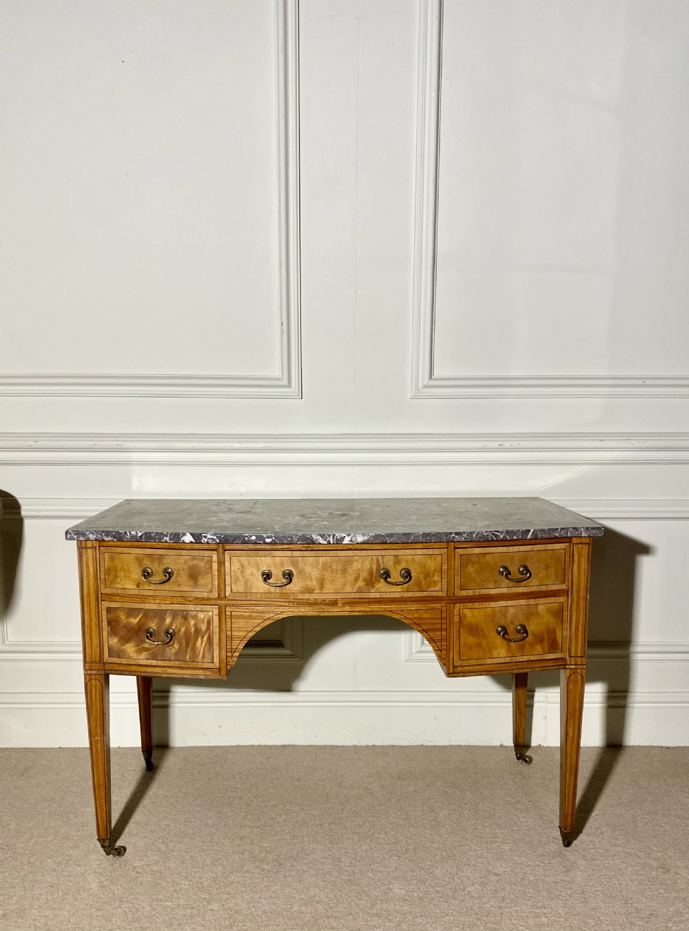 A 19th-century satinwood dressing table featuring a grey marble top, five drawers with brass drop handles, a central arch, and tapered legs on castors. View III