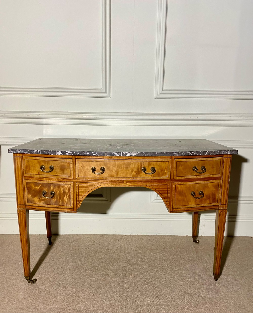 A 19th-century satinwood dressing table featuring a grey marble top, five drawers with brass drop handles, a central arch, and tapered legs on castors. View V