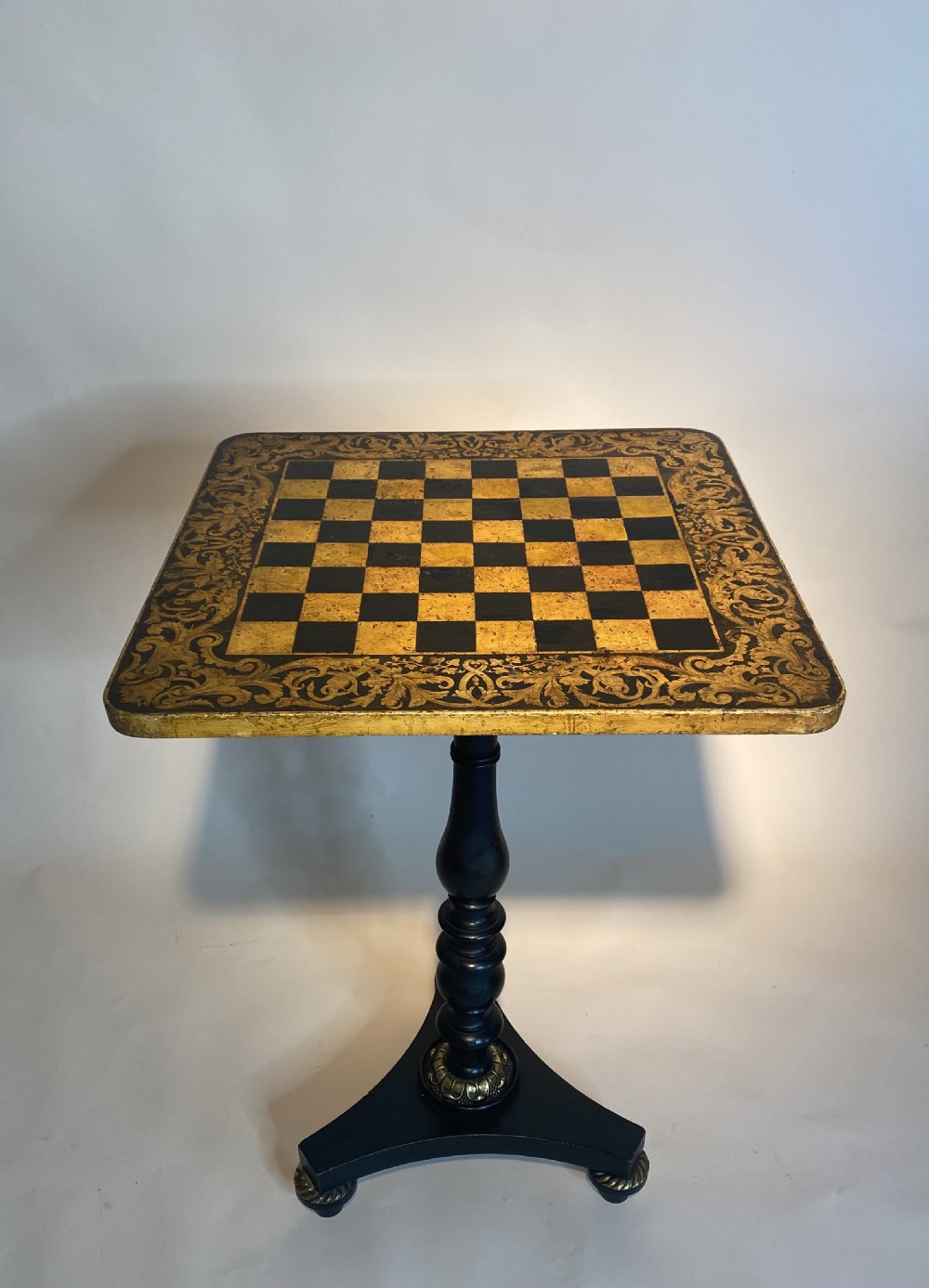A square Regency ebonised chess table featuring detailed foliate penwork framing a central chessboard top, supported by a turned central pedestal and tripod base.