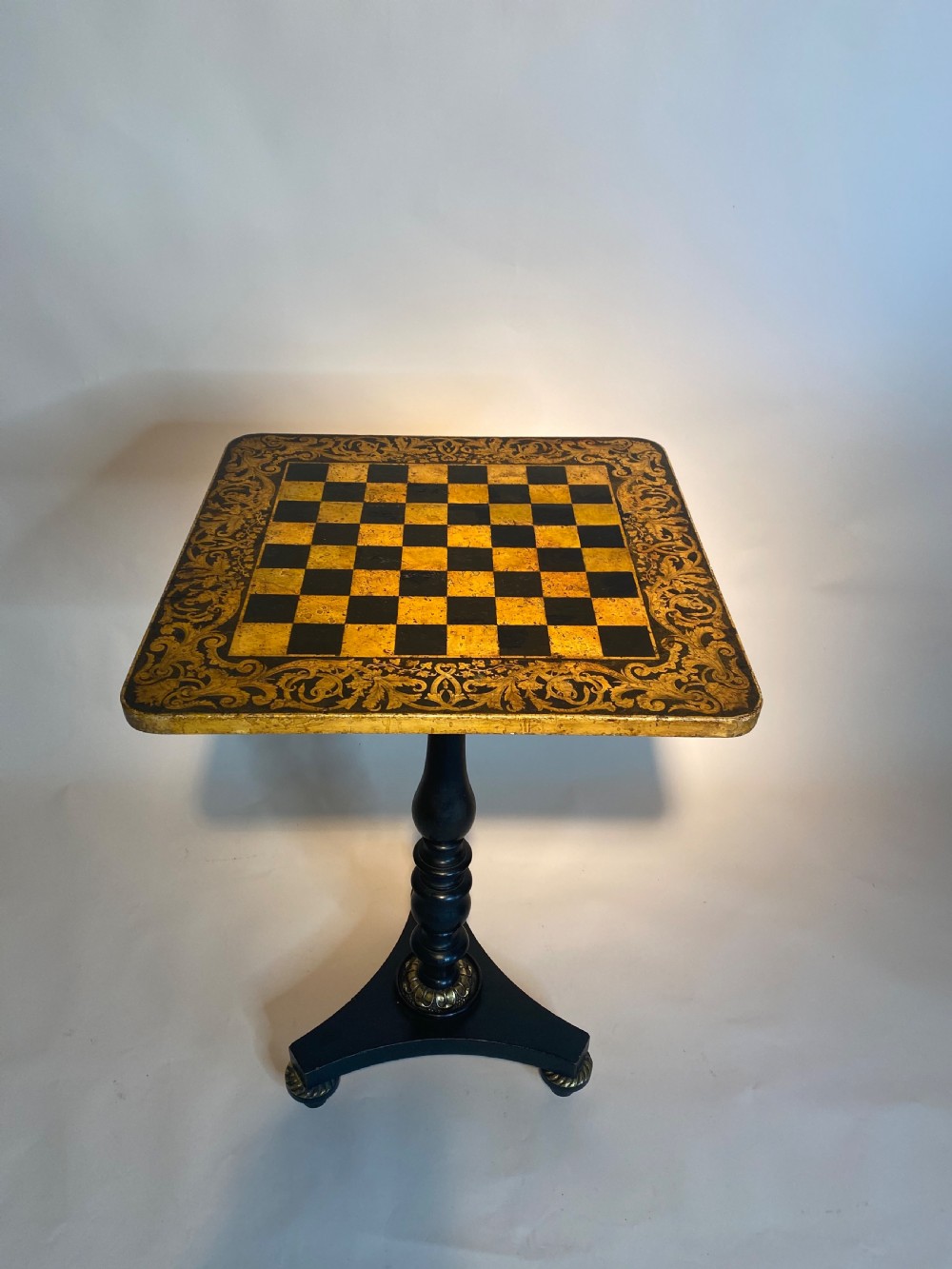 A square Regency ebonised chess table featuring detailed foliate penwork framing a central chessboard top, supported by a turned central pedestal and tripod base. View II