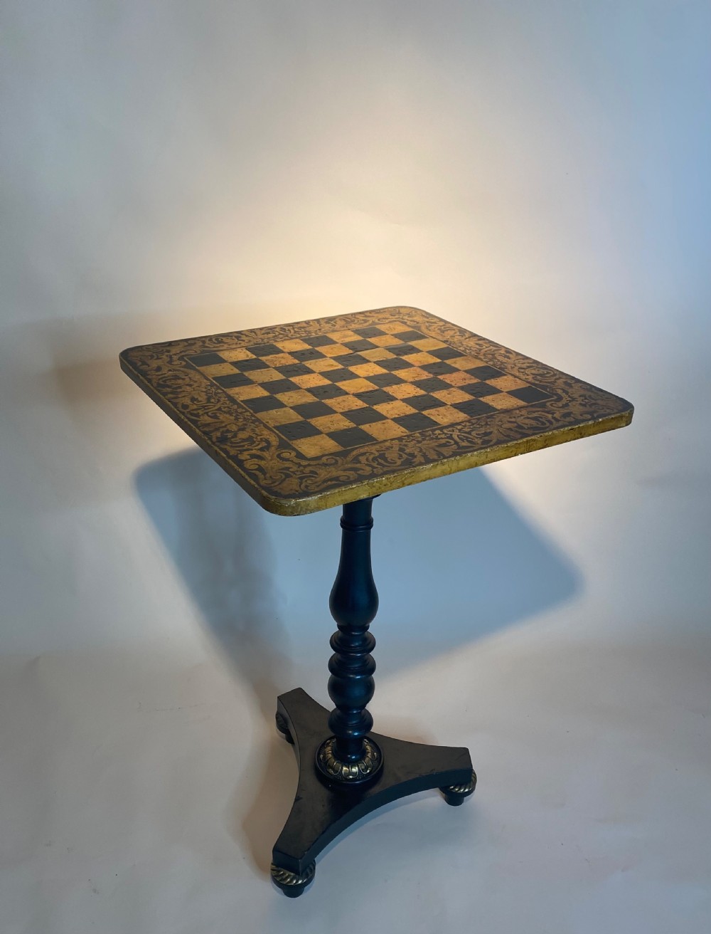 A square Regency ebonised chess table featuring detailed foliate penwork framing a central chessboard top, supported by a turned central pedestal and tripod base. View V