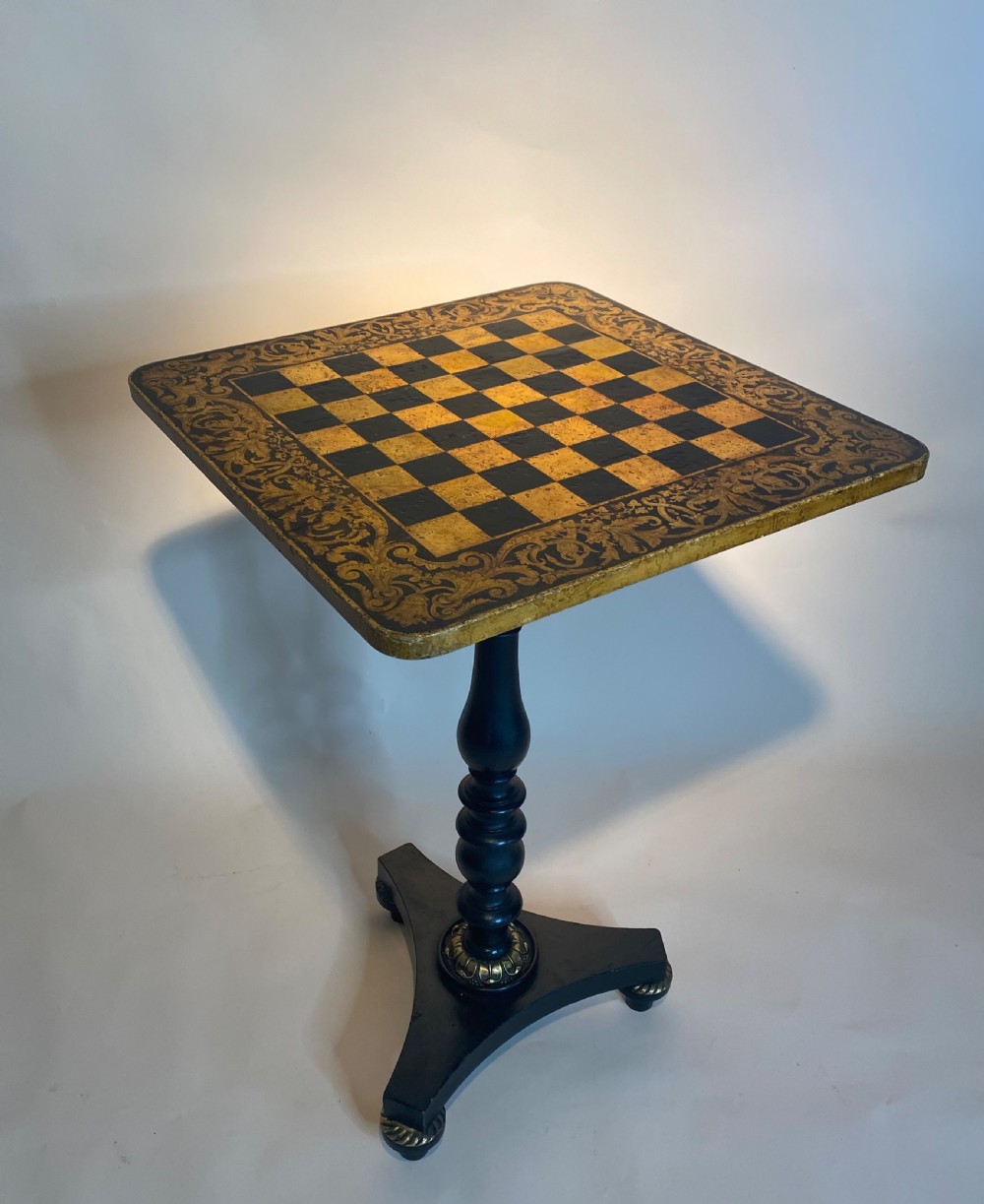 A square Regency ebonised chess table featuring detailed foliate penwork framing a central chessboard top, supported by a turned central pedestal and tripod base. View VI