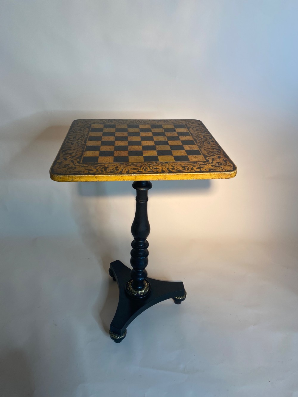 A square Regency ebonised chess table featuring detailed foliate penwork framing a central chessboard top, supported by a turned central pedestal and tripod base. View VIII