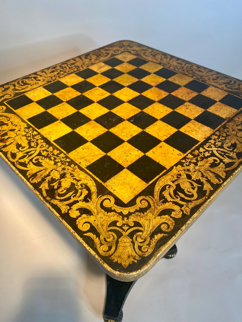 A square Regency ebonised chess table featuring detailed foliate penwork framing a central chessboard top, supported by a turned central pedestal and tripod base. View XIII