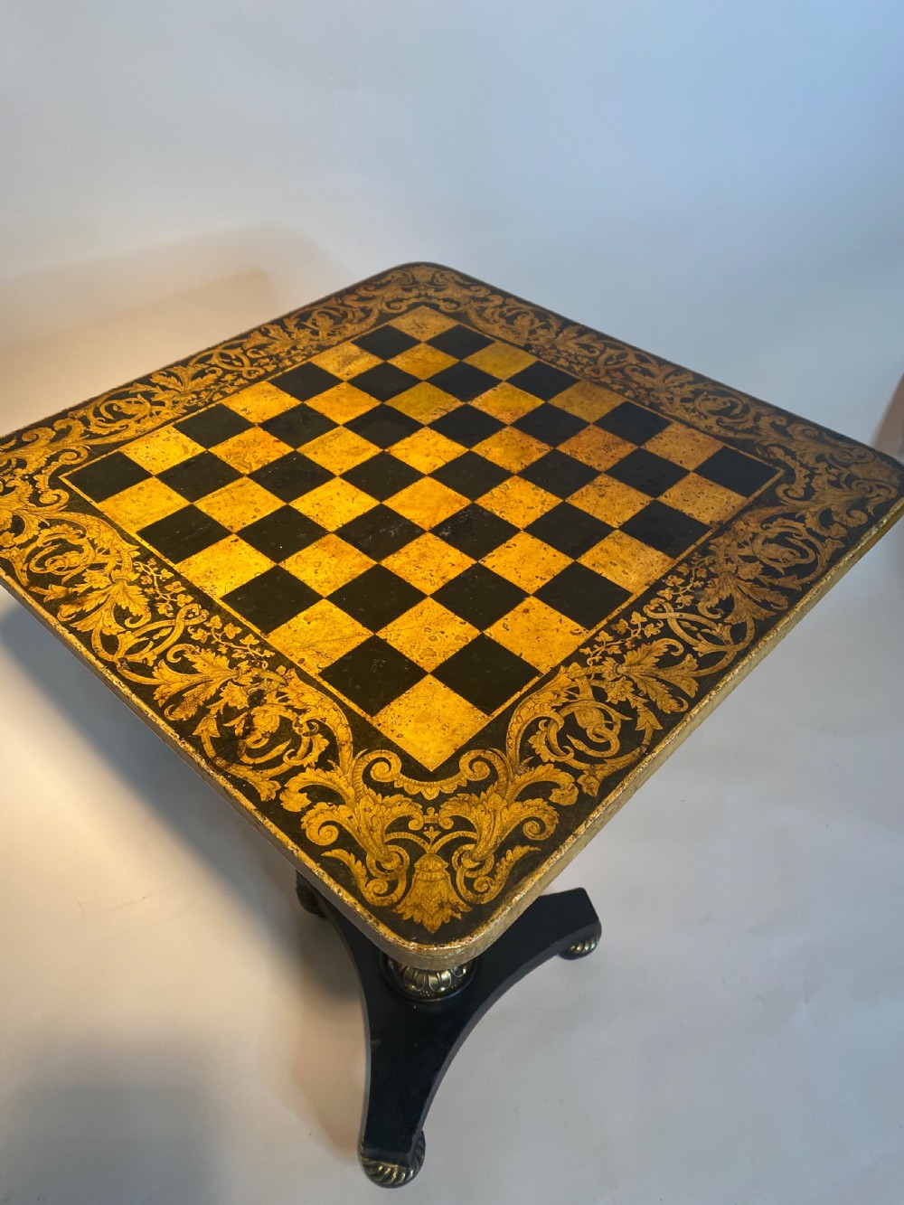 A square Regency ebonised chess table featuring detailed foliate penwork framing a central chessboard top, supported by a turned central pedestal and tripod base. View XIV