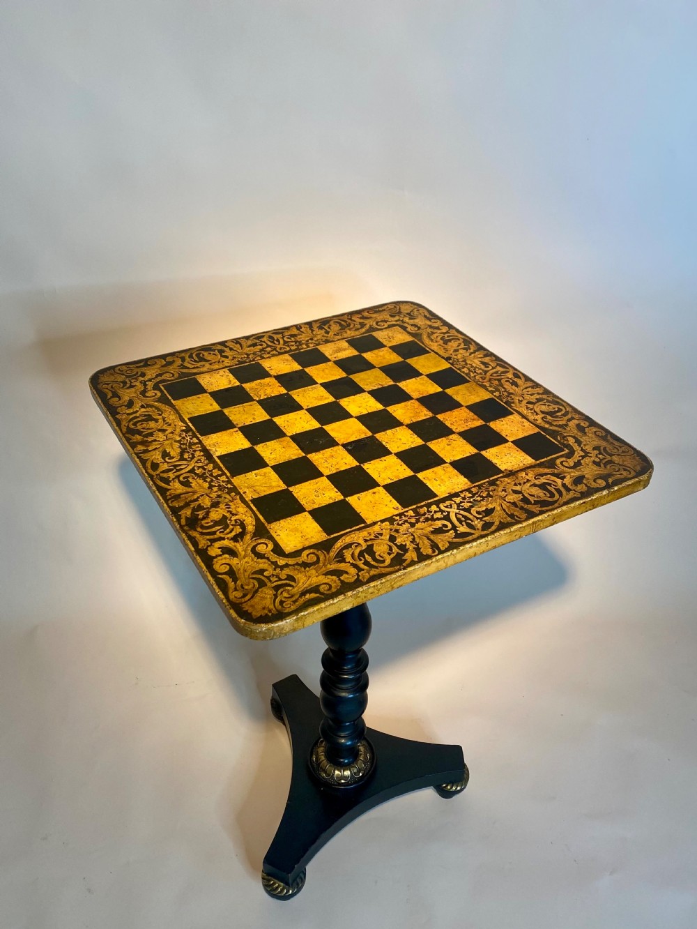 A square Regency ebonised chess table featuring detailed foliate penwork framing a central chessboard top, supported by a turned central pedestal and tripod base. View XV