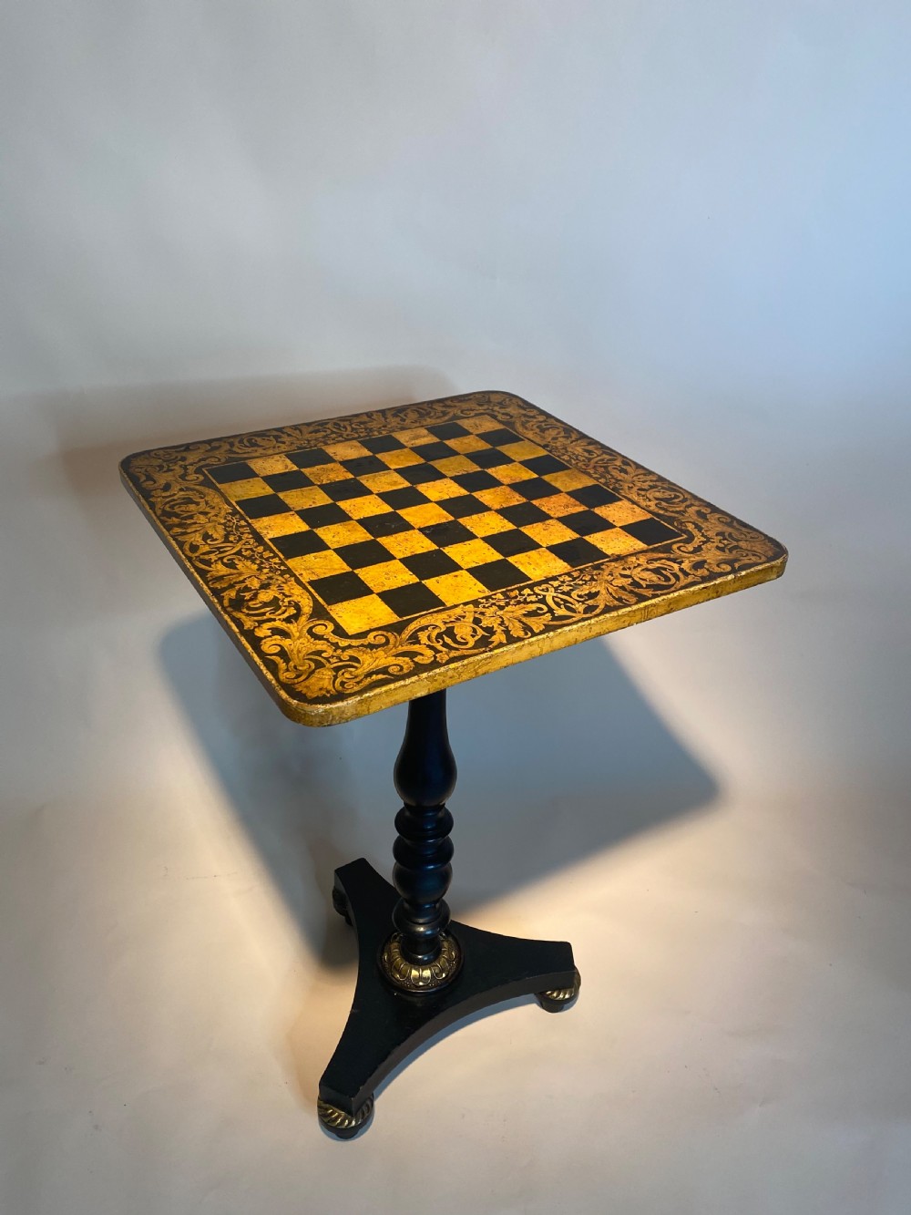A square Regency ebonised chess table featuring detailed foliate penwork framing a central chessboard top, supported by a turned central pedestal and tripod base. View XVI