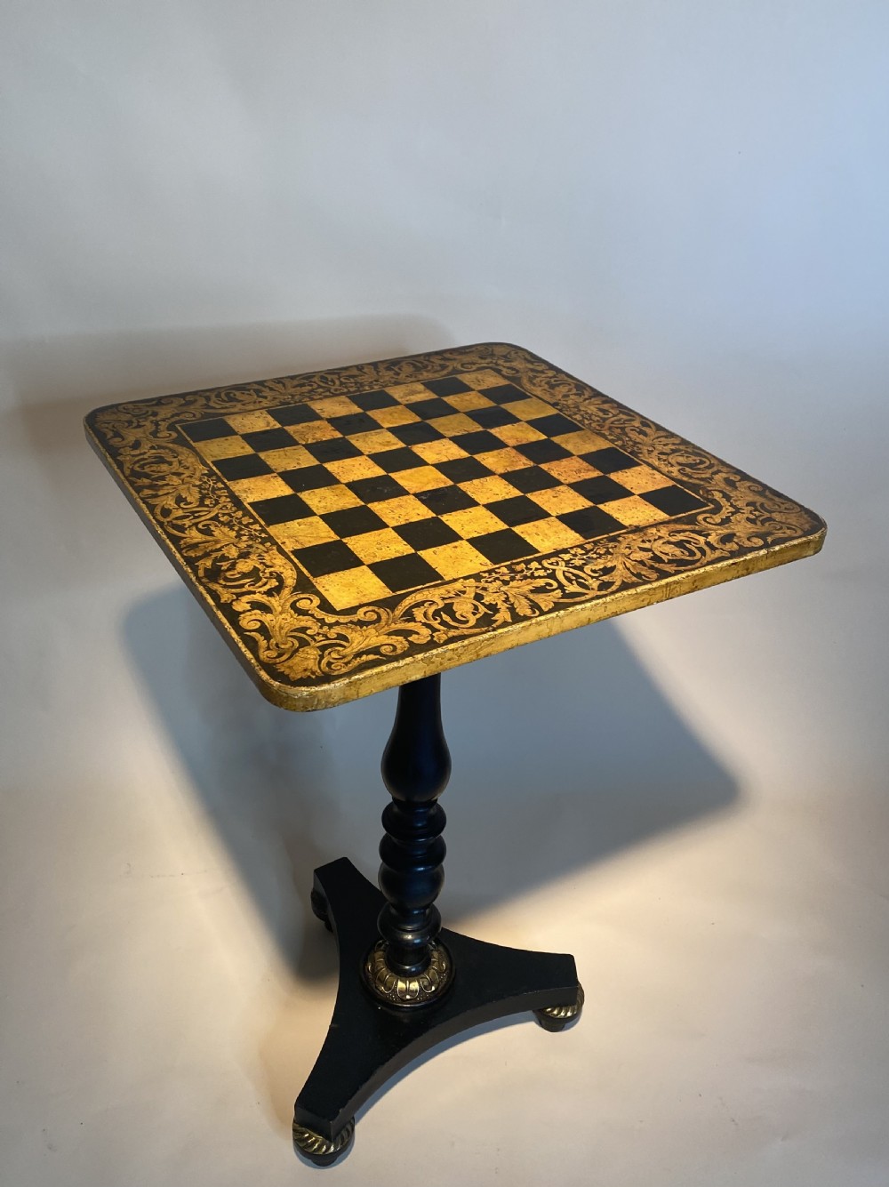 A square Regency ebonised chess table featuring detailed foliate penwork framing a central chessboard top, supported by a turned central pedestal and tripod base. View XVII
