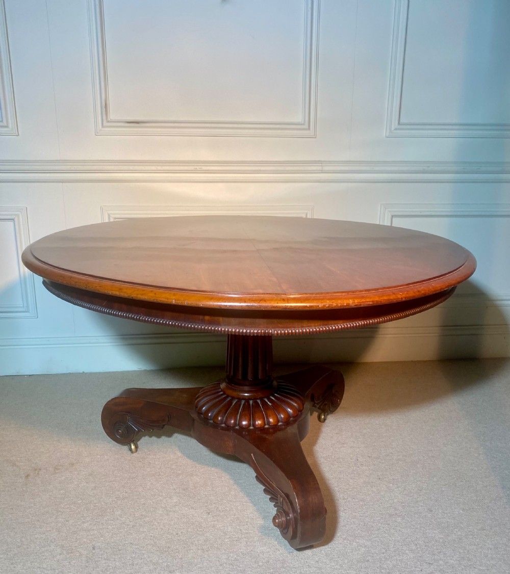 An antique Regency mahogany oval breakfast table featuring a carved fluted pedestal support and scroll feet with brass castors.