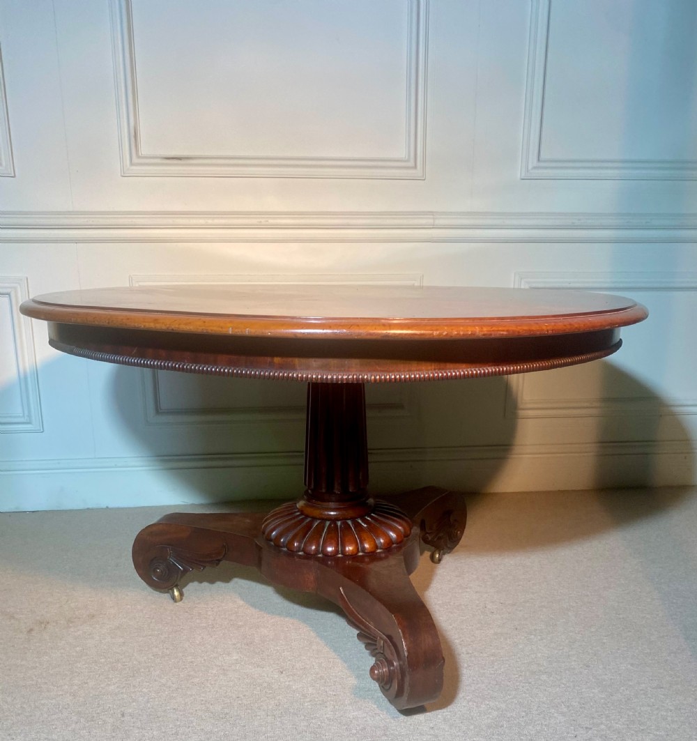 An antique Regency mahogany oval breakfast table featuring a carved fluted pedestal support and scroll feet with brass castors. View II