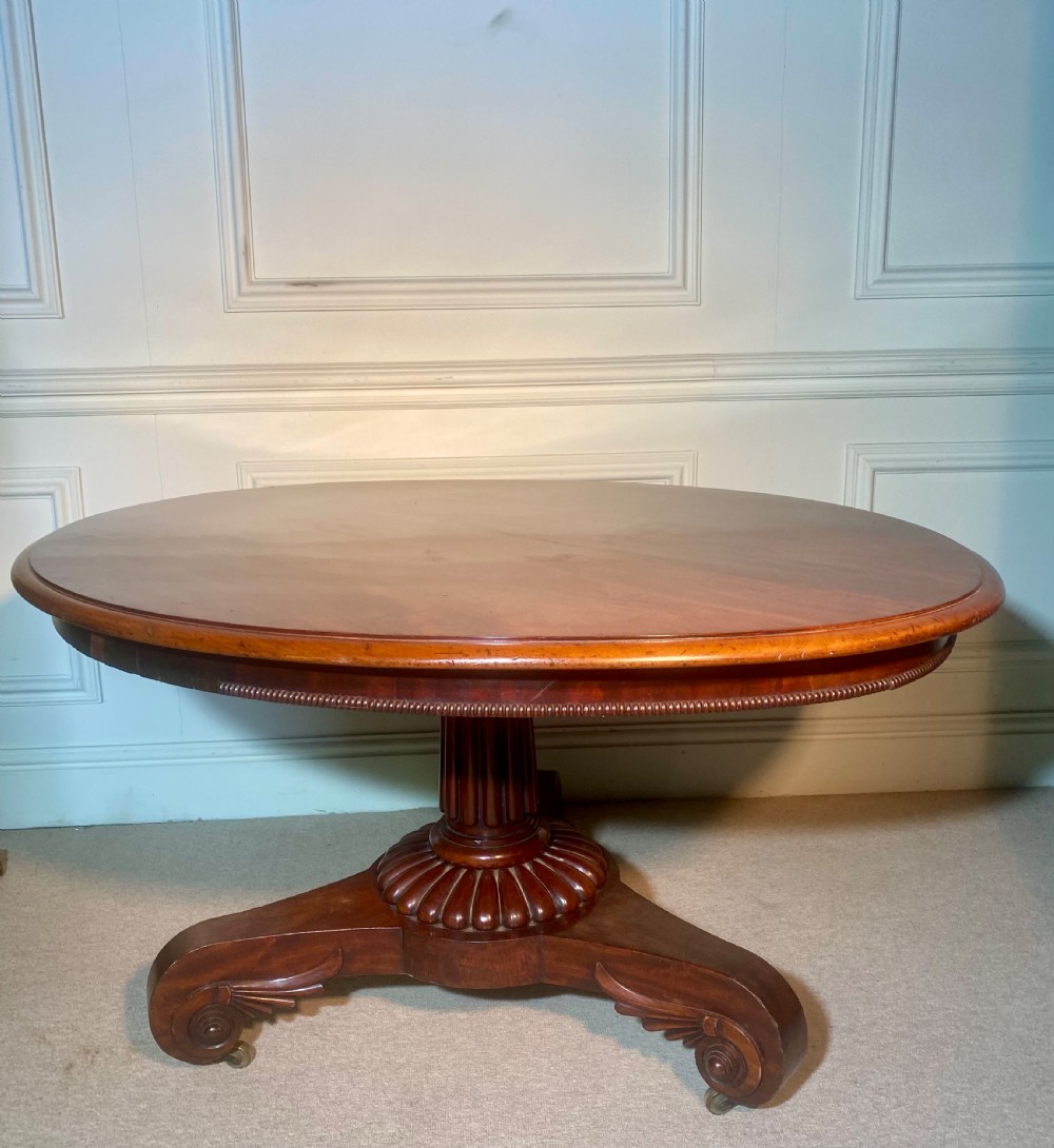 An antique Regency mahogany oval breakfast table featuring a carved fluted pedestal support and scroll feet with brass castors. View V