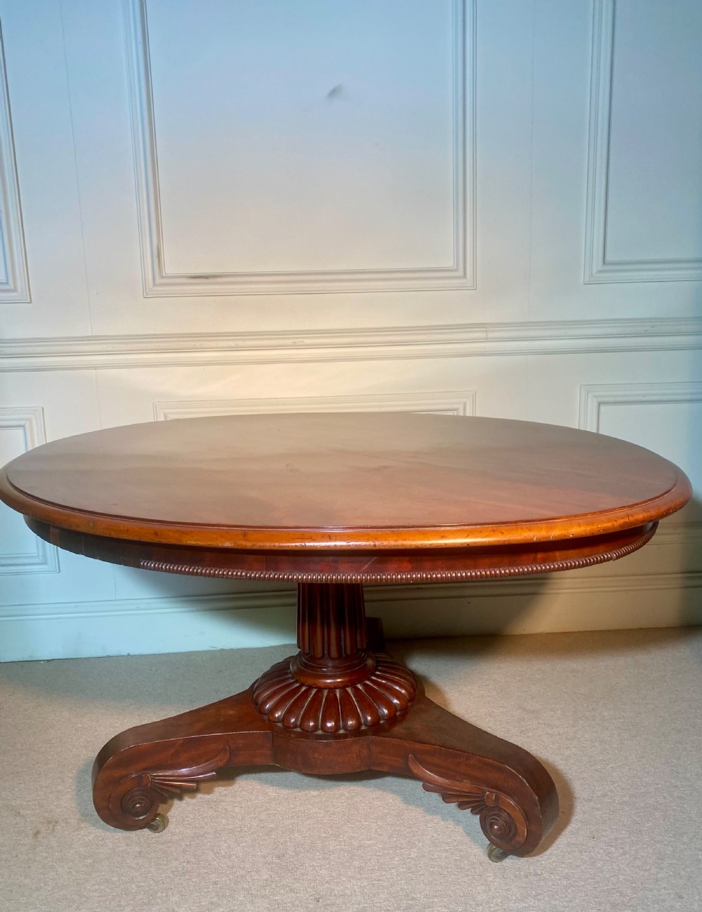 An antique Regency mahogany oval breakfast table featuring a carved fluted pedestal support and scroll feet with brass castors. View VI