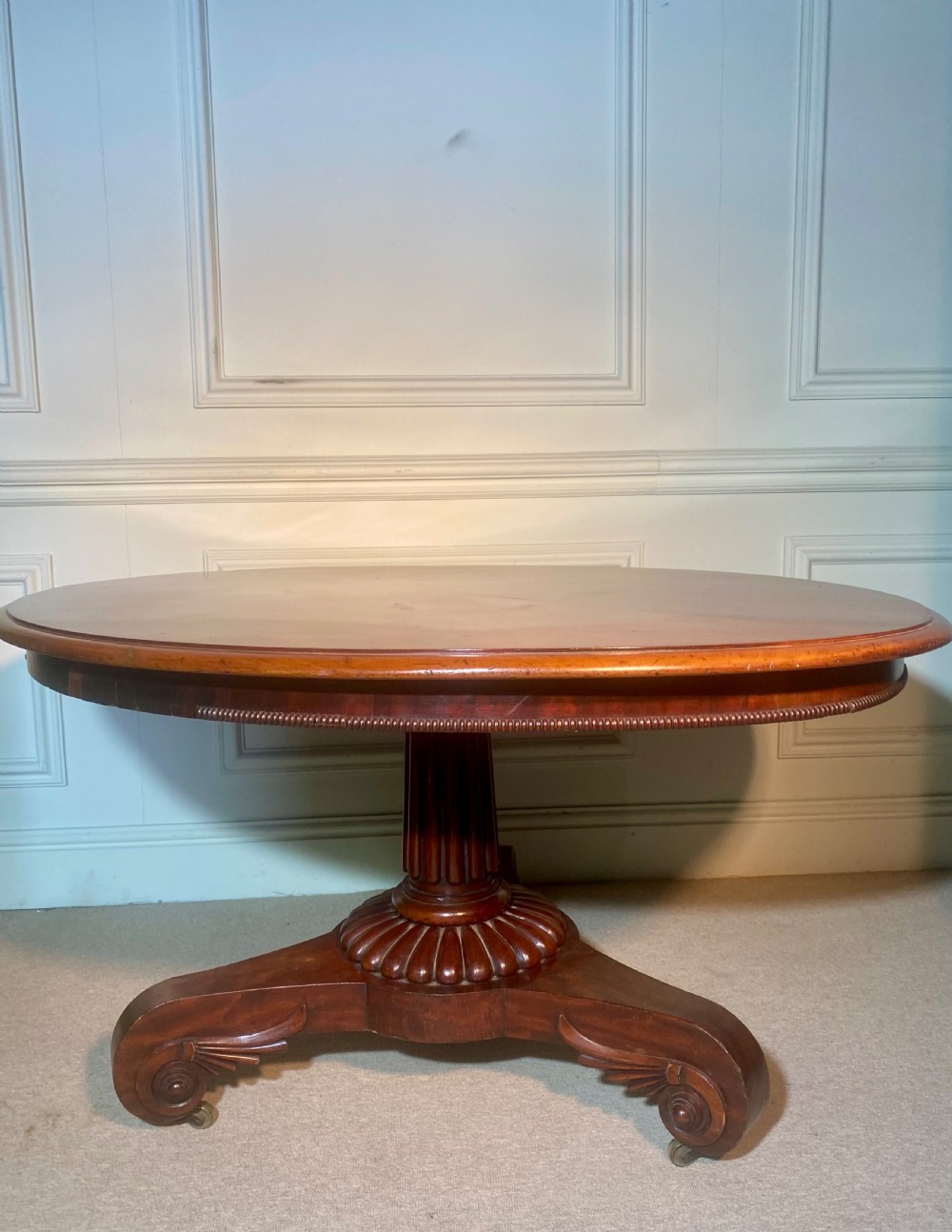 An antique Regency mahogany oval breakfast table featuring a carved fluted pedestal support and scroll feet with brass castors. View VII