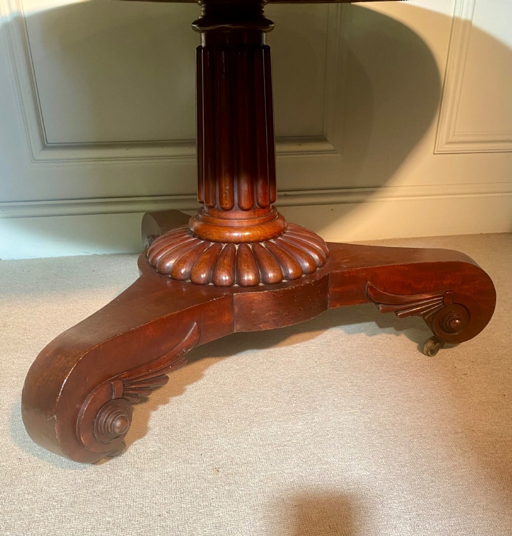 An antique Regency mahogany oval breakfast table featuring a carved fluted pedestal support and scroll feet with brass castors. View X