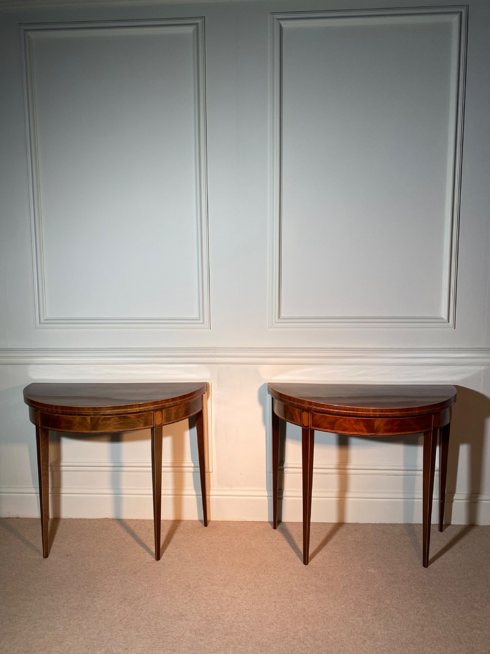 A pair of 18th-century George III mahogany demi-lune card tables with inlaid friezes and square tapered legs positioned against a white panelled wall.