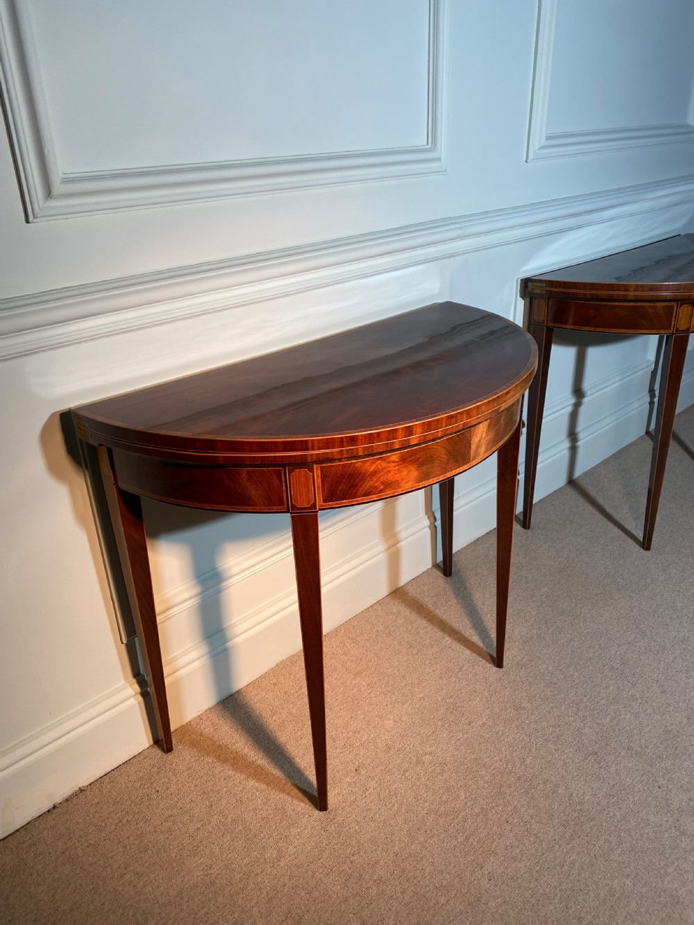 A pair of 18th-century George III mahogany demi-lune card tables with inlaid friezes and square tapered legs positioned against a white panelled wall. View VI