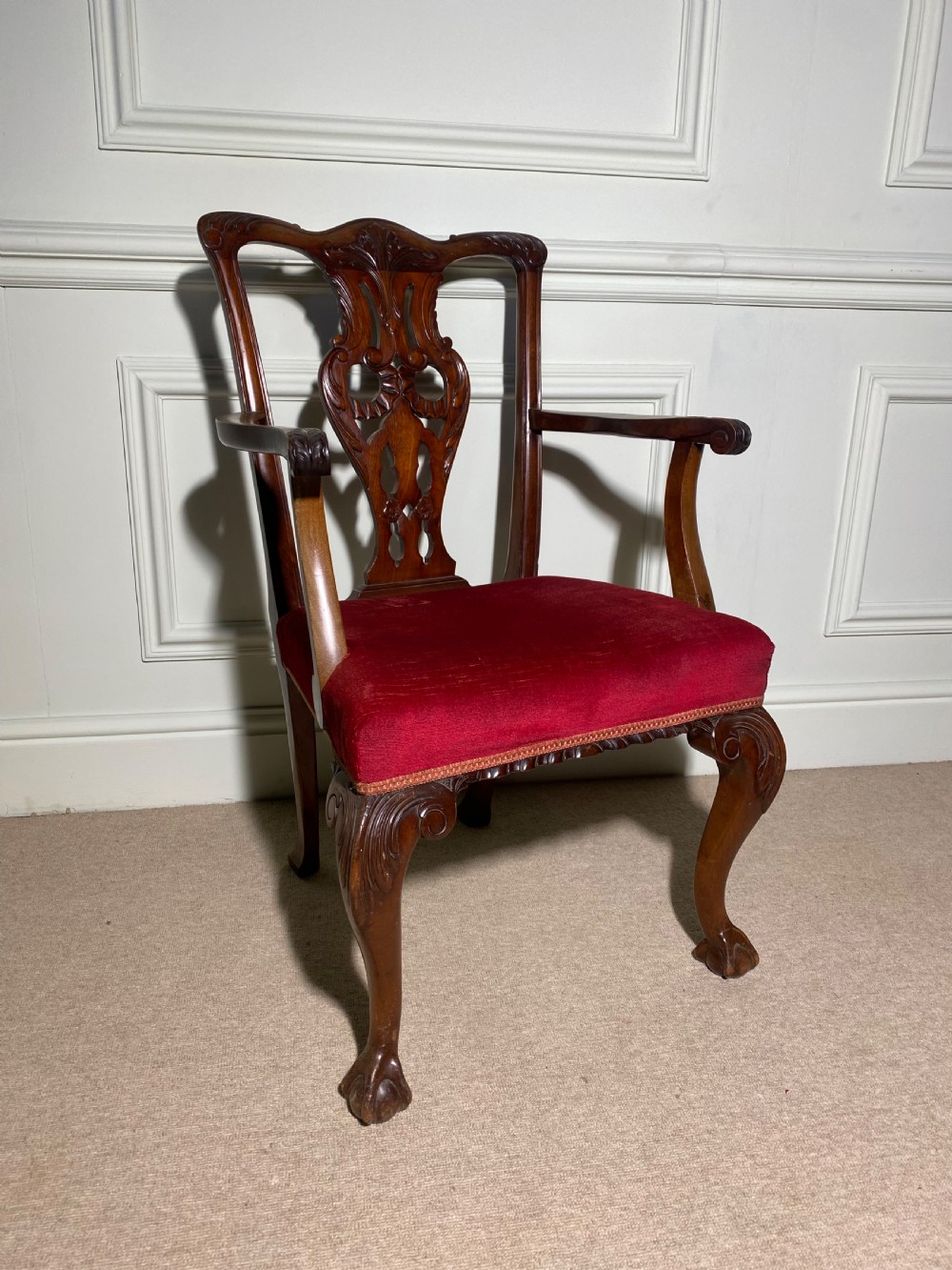 A Chippendale style carved mahogany carver chair featuring a pierced splat back, shaped scroll arms, red velvet seat cushion, and cabriole legs with ball and claw feet.