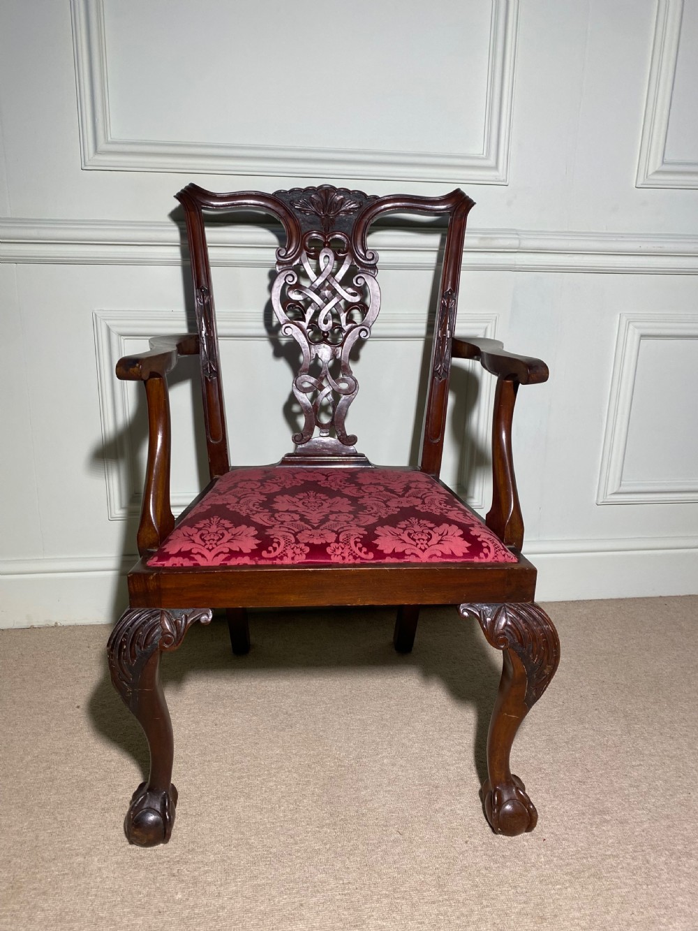 A Chippendale style mahogany carver armchair featuring an intricately carved pierced splat back, red patterned damask upholstery, and cabriole legs with ball and claw feet.