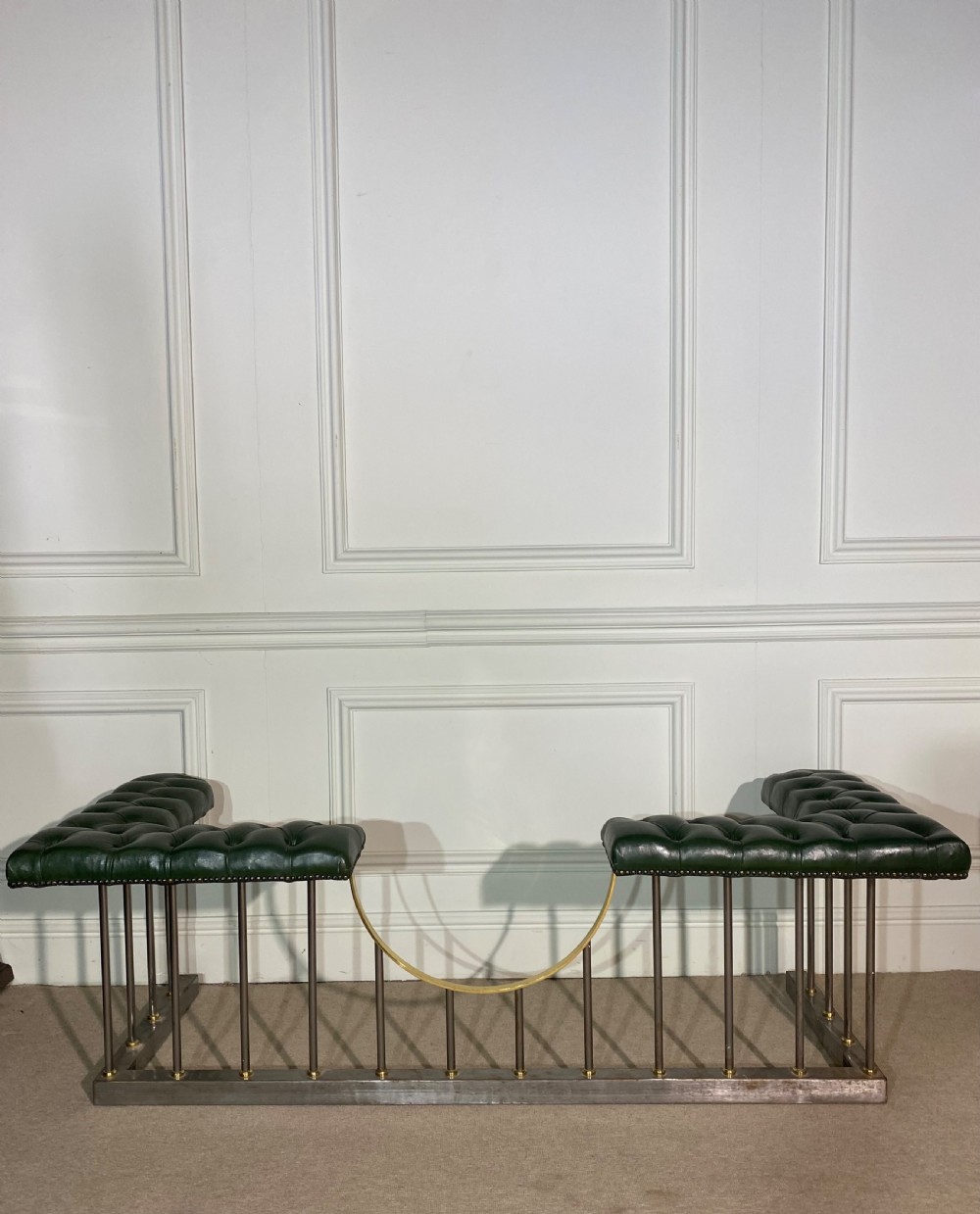 A metal fireplace club fender with vertical steel uprights, brass accents, a central curved dip, and two corner seats upholstered in button-tufted dark green leather with brass studding.