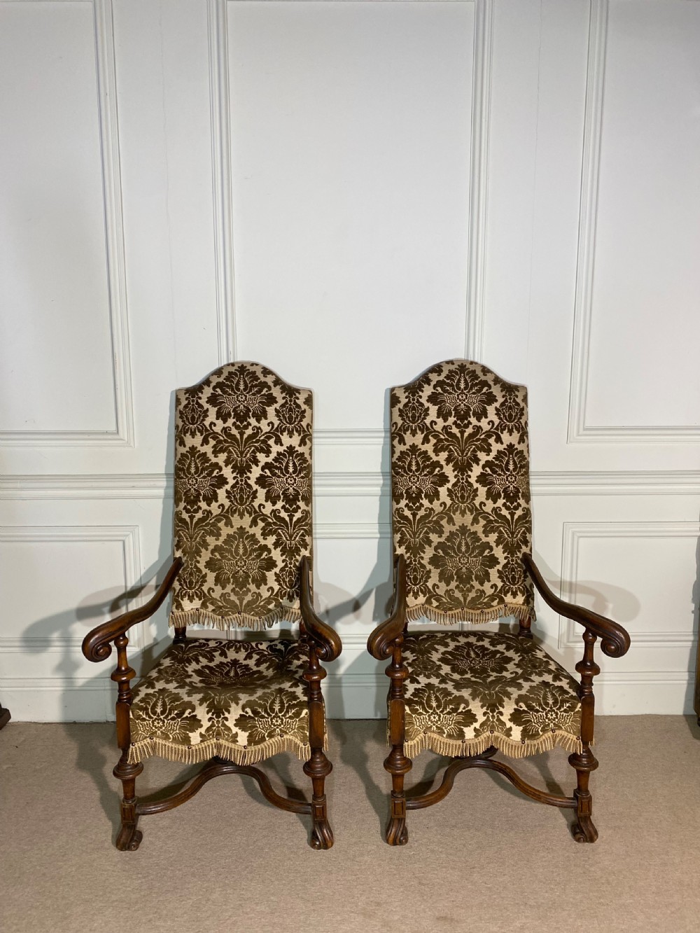 A pair of high-back Carolean style armchairs upholstered in patterned brown velvet with fringe trim and carved timber frames.