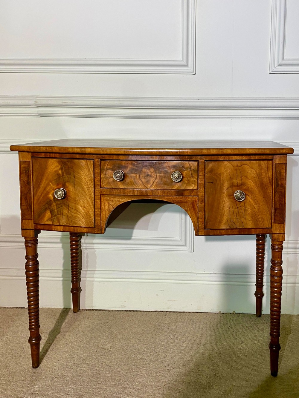 A George III bow-fronted mahogany dressing table featuring three drawers with round brass knobs, an arched central kneehole, and four turned legs.
