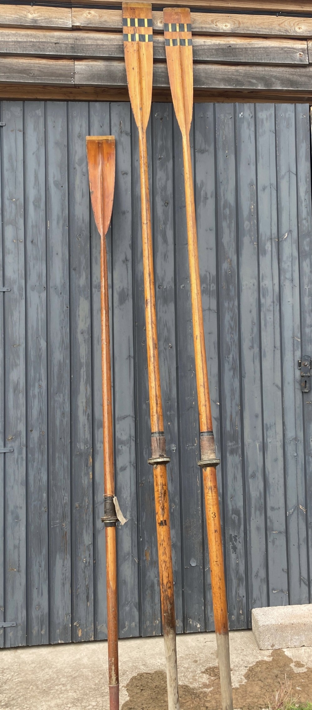 A pair of tall antique wooden rowing oars featuring long timber shafts, stitched leather collars, and painted geometric band markings on the blades.