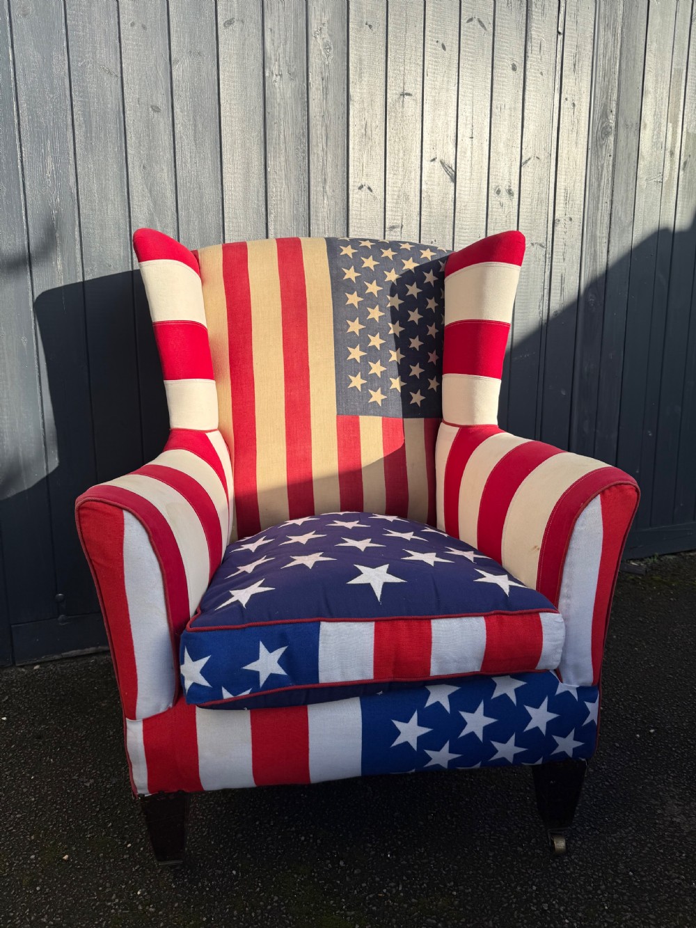 A Victorian style wingback armchair upholstered in an American stars and stripes flag pattern with turned dark wood front legs on brass castors. View III